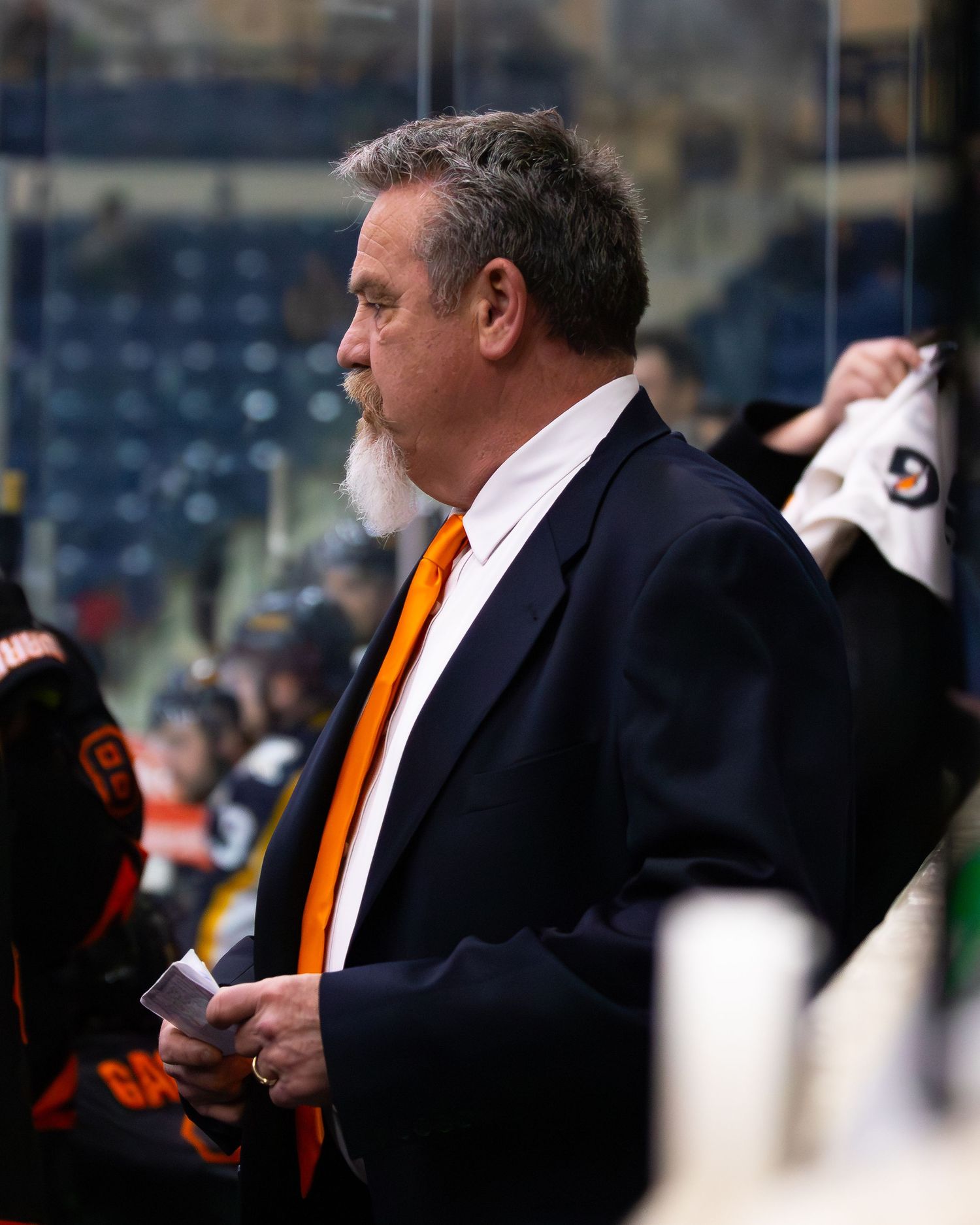 Senior hockey coach in black blazer with orange tie stands behind bench at ice rink.