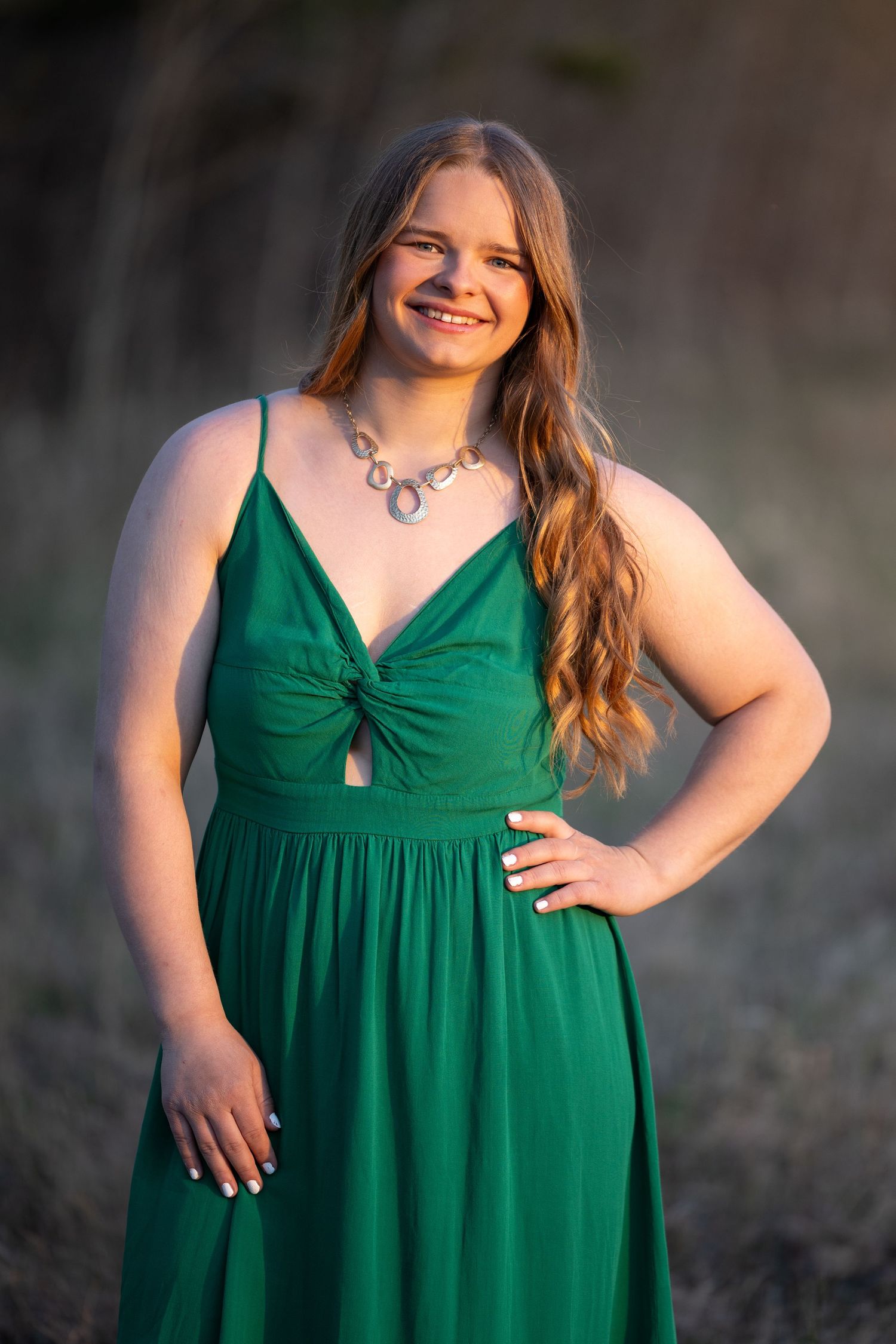 Young woman smiling in elegant green spaghetti strap dress with silver necklace, posing outdoors in golden hour light.
