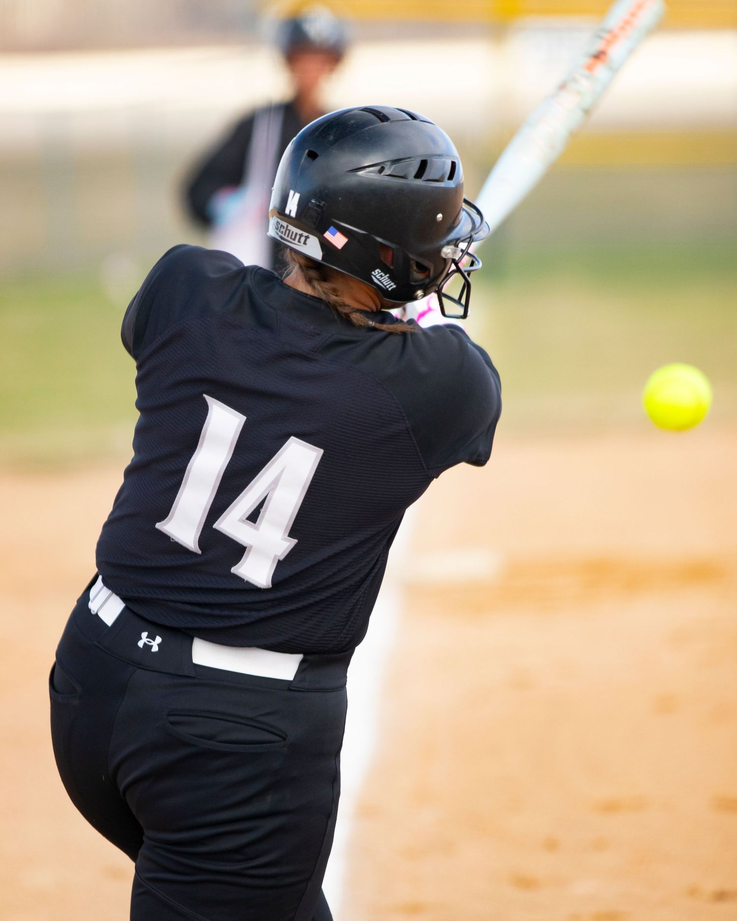 Softball batter in black jersey number 14 making contact with yellow ball during game.