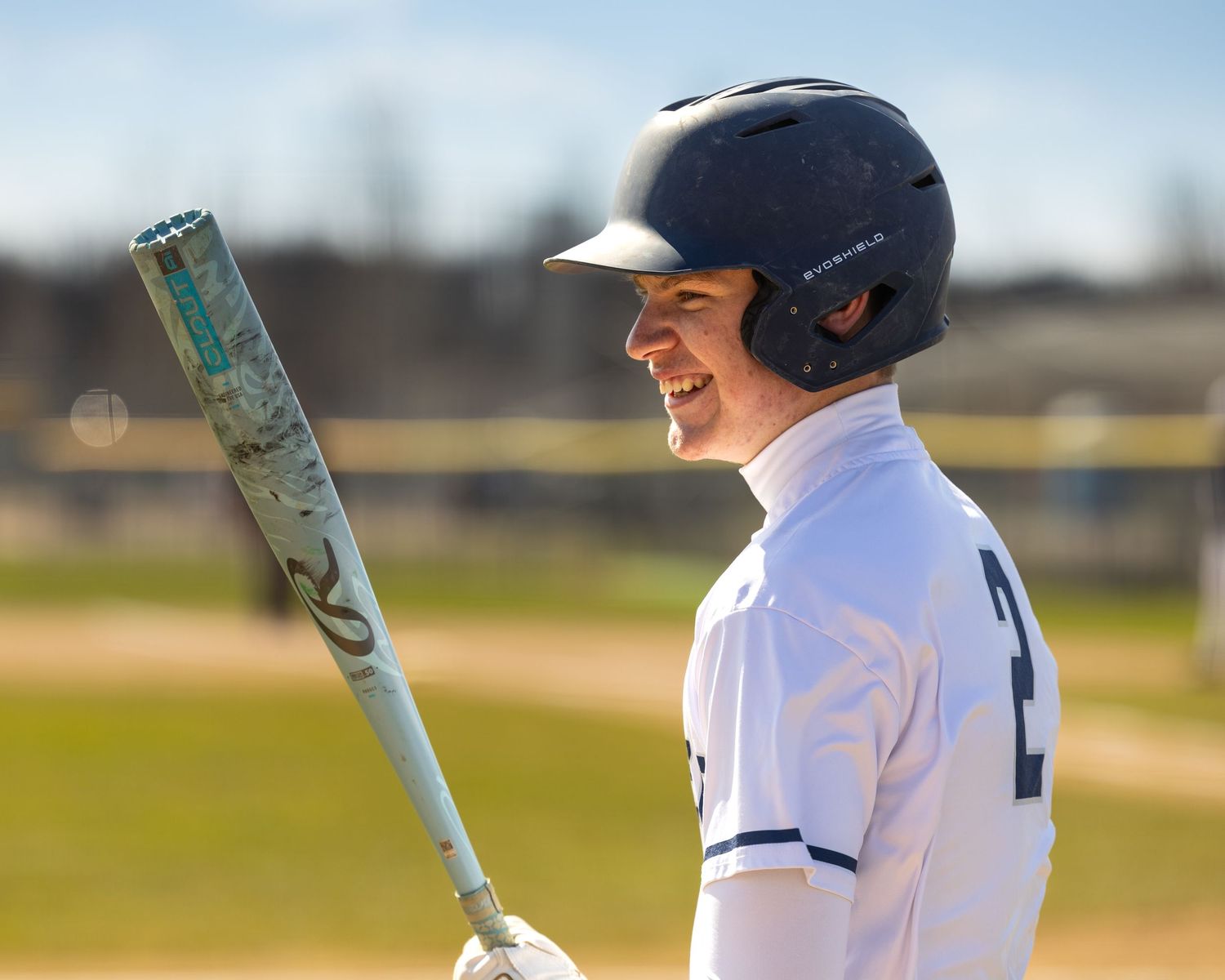 Smiling baseball player in white uniform and black helmet holding a bat on a sunny outdoor field.
