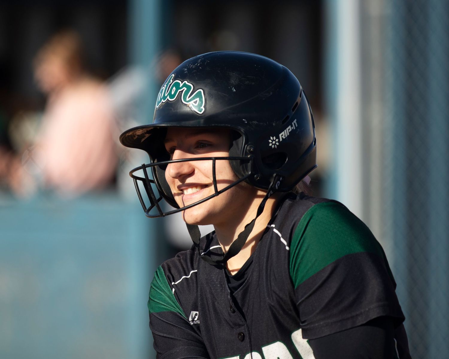 Smiling softball player wearing a black batting helmet with face guard in green and black team uniform.