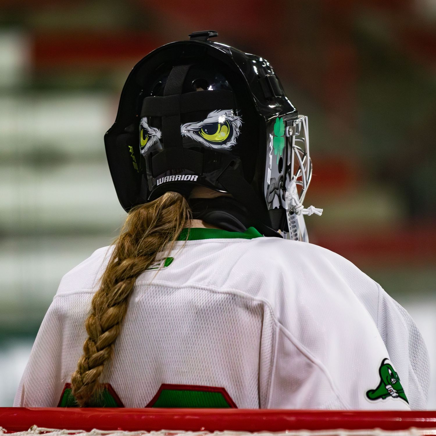 Female hockey goalie with braided hair and decorated black helmet faces away.