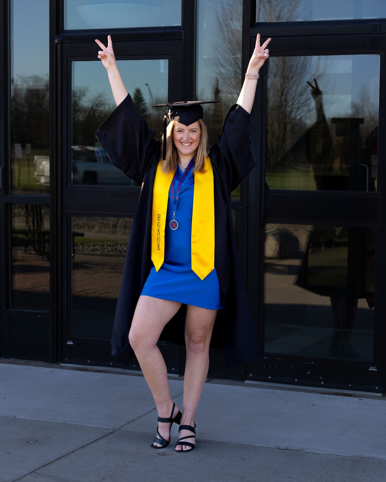 A smiling graduate in black cap and gown with yellow stole poses with arms raised in victory signs outside a building.