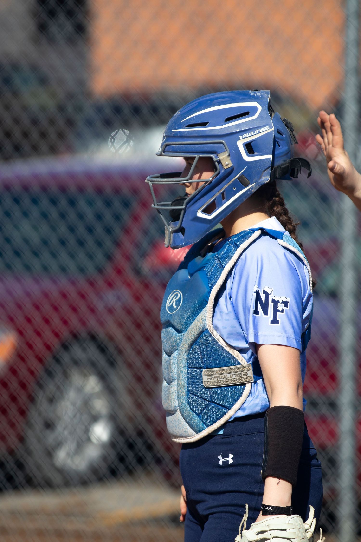 Female softball catcher in blue NF uniform and Rawlings gear stands ready during a sunny outdoor game.