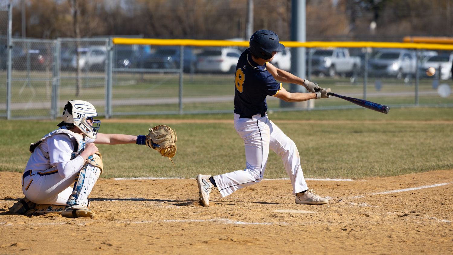 Youth baseball batter swinging at pitch with catcher crouching behind home plate on sunny day.