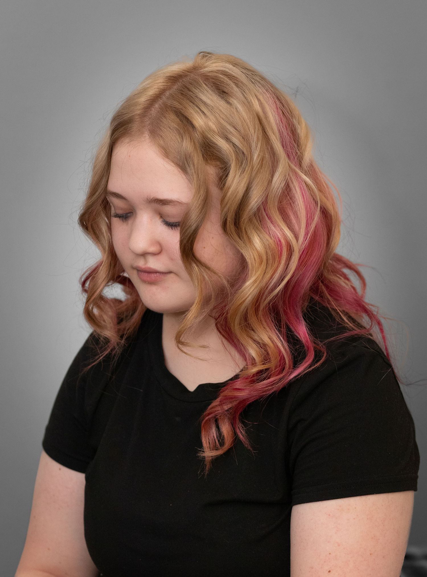 Young woman with wavy blonde hair featuring pink and red highlights, wearing a black top against a gray background.