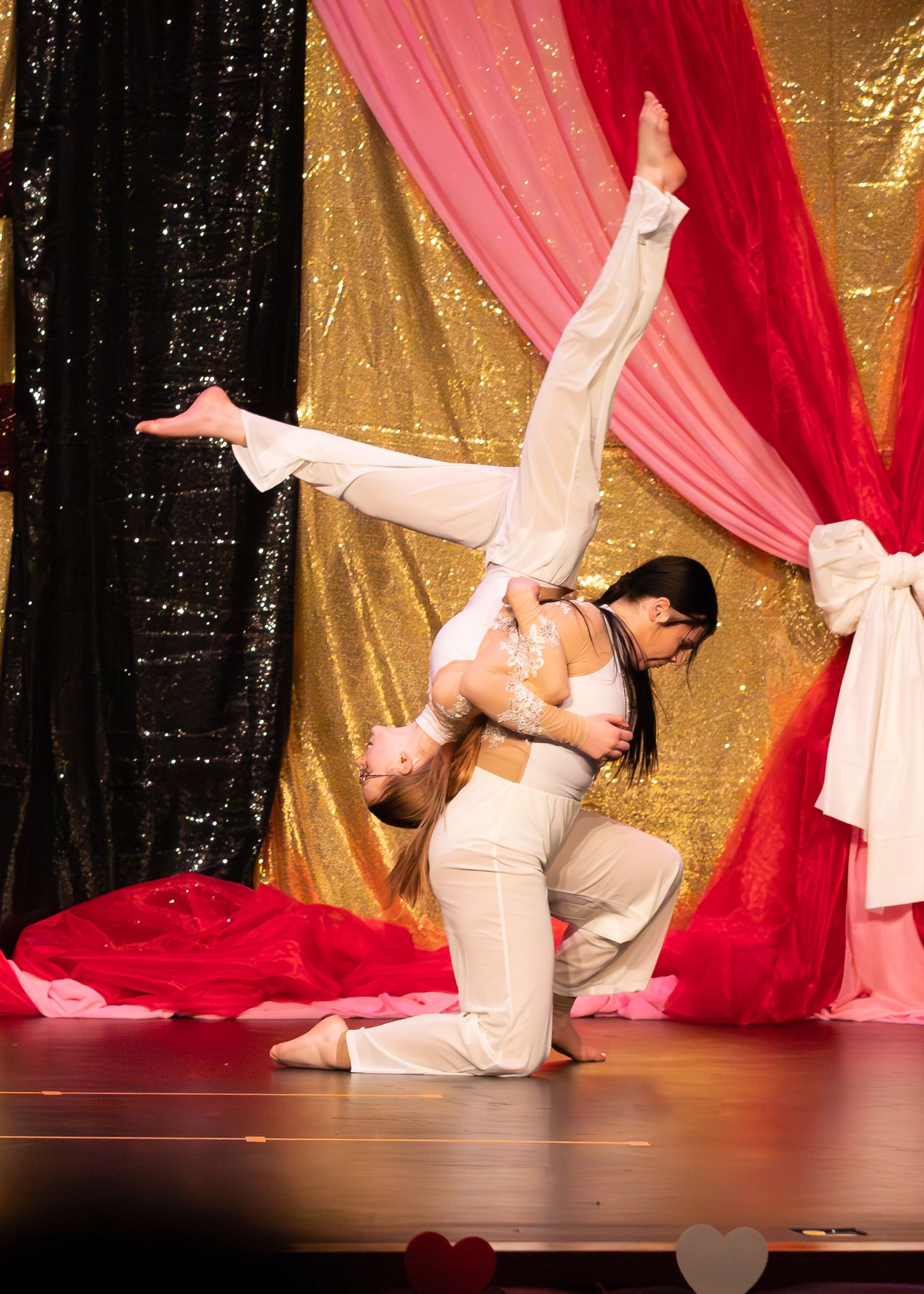 Two dancers in white perform an acrobatic lift on stage with gold and pink sequined curtain backdrop.