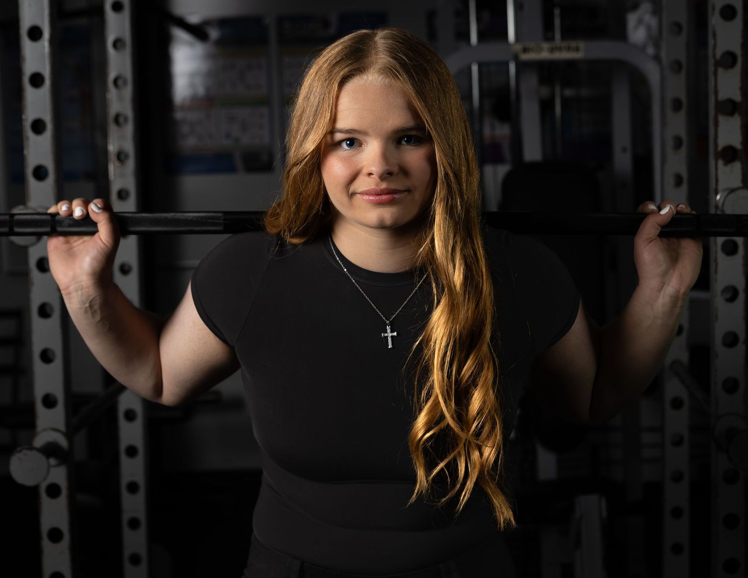 Young woman with long red hair poses with a barbell on her shoulders in a gym, wearing a cross necklace.