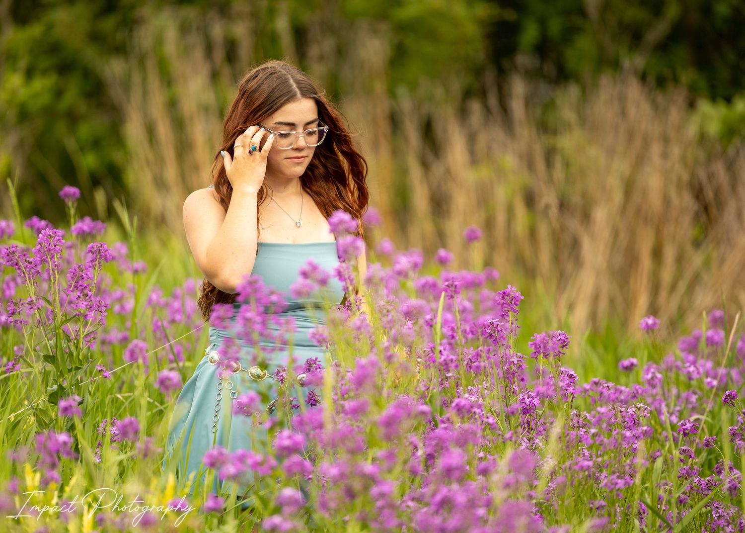 Young woman in blue dress adjusting glasses among vibrant purple wildflowers in a lush green meadow.