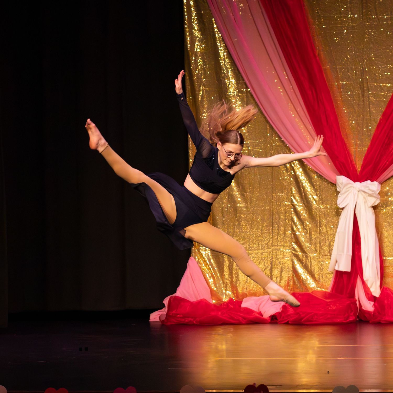 Dancer in black outfit executes a graceful aerial leap on stage with gold sequin and red drape backdrop.
