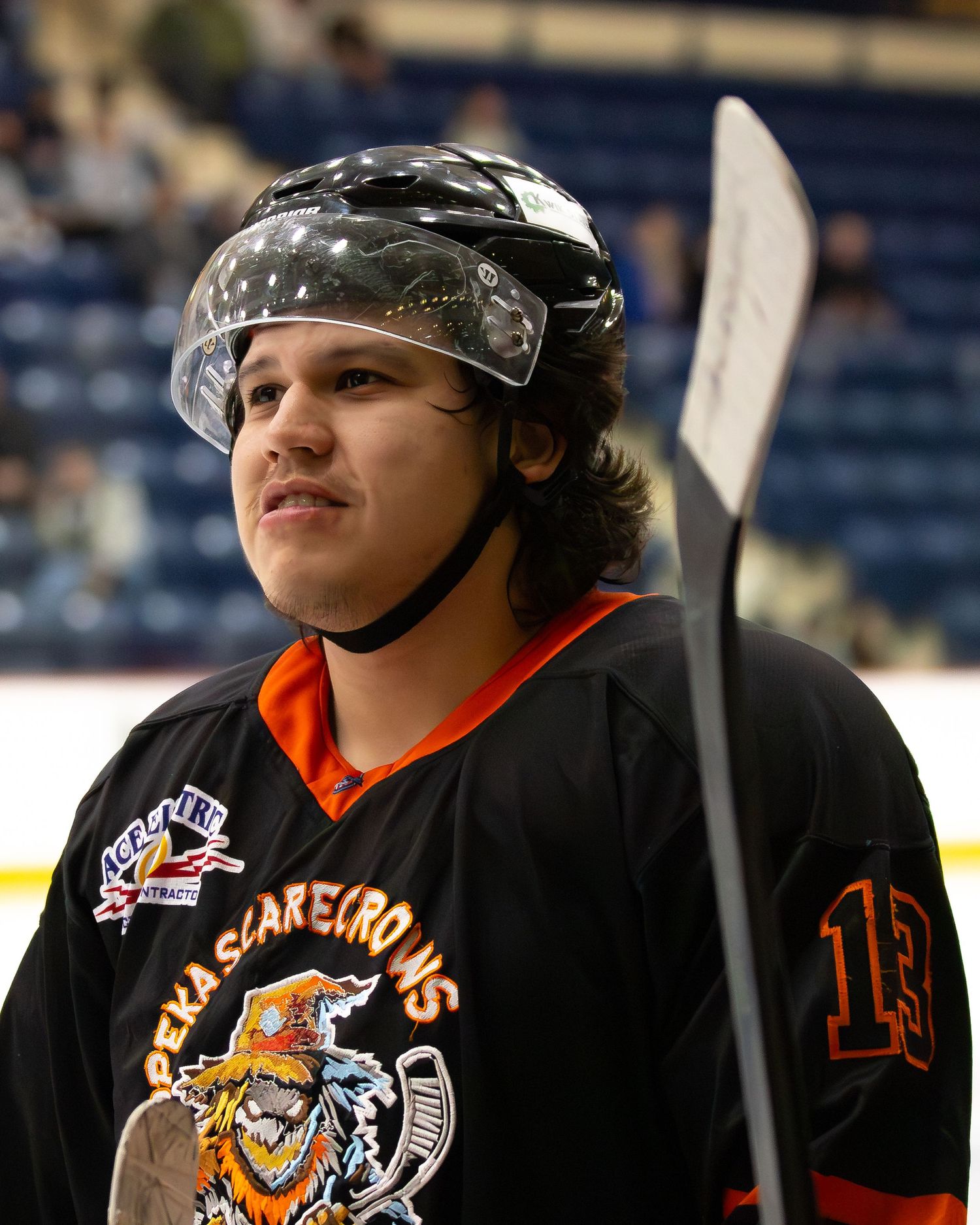 Young hockey player in black Scarecrows jersey wearing helmet with visor holds stick on ice rink.
