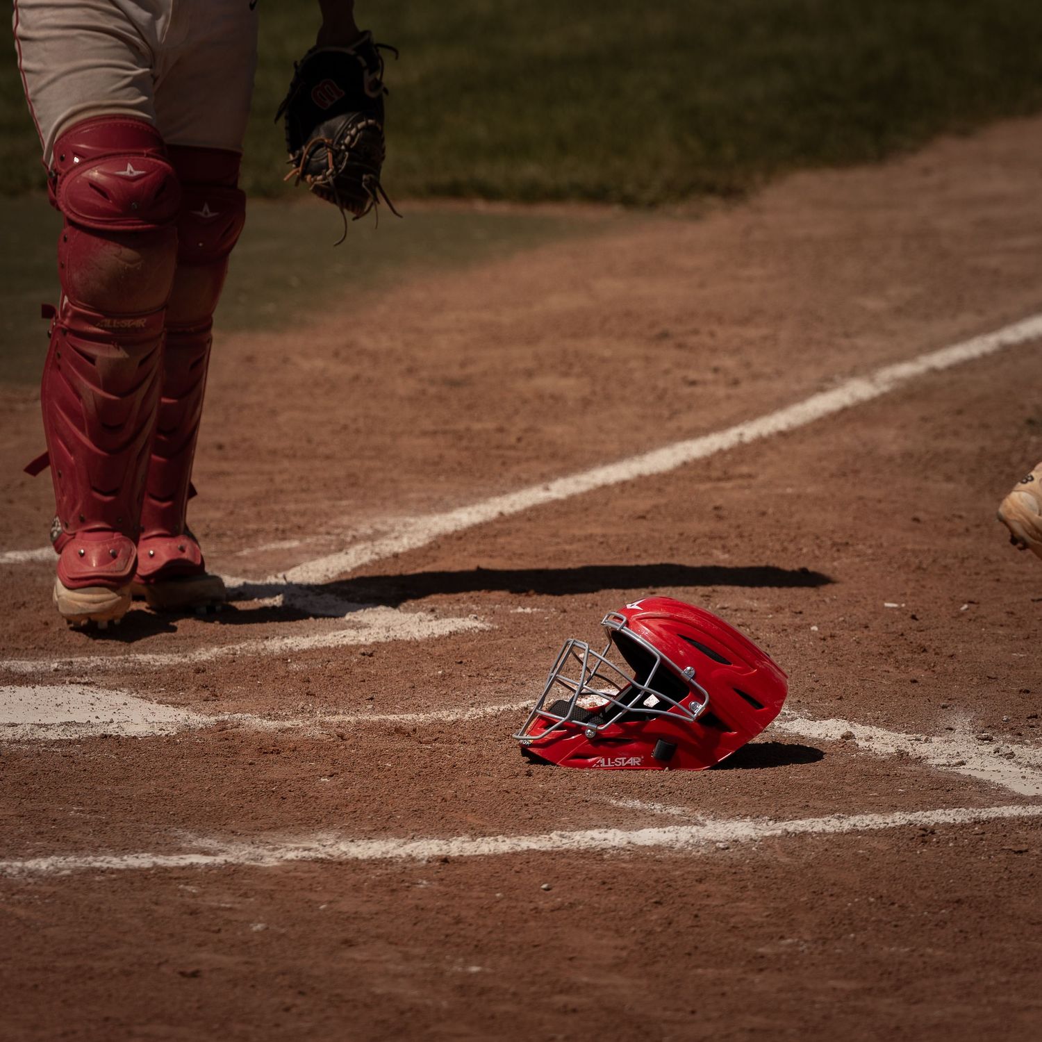 Red baseball helmet on home plate with catcher in gear standing nearby on a sunny dirt baseball field.