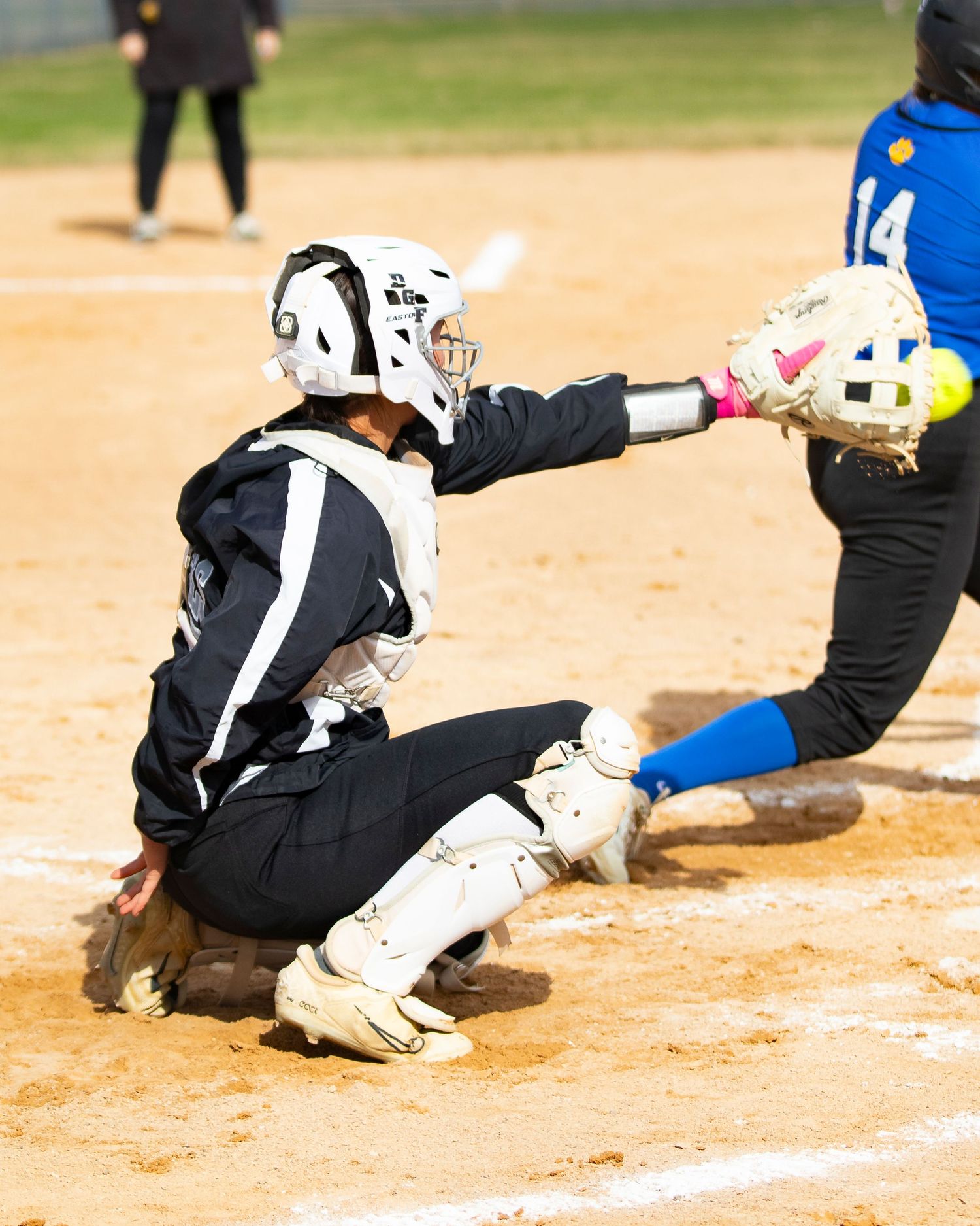 Softball catcher in white helmet and gear crouching at home plate during a game, reaching to tag out a runner.