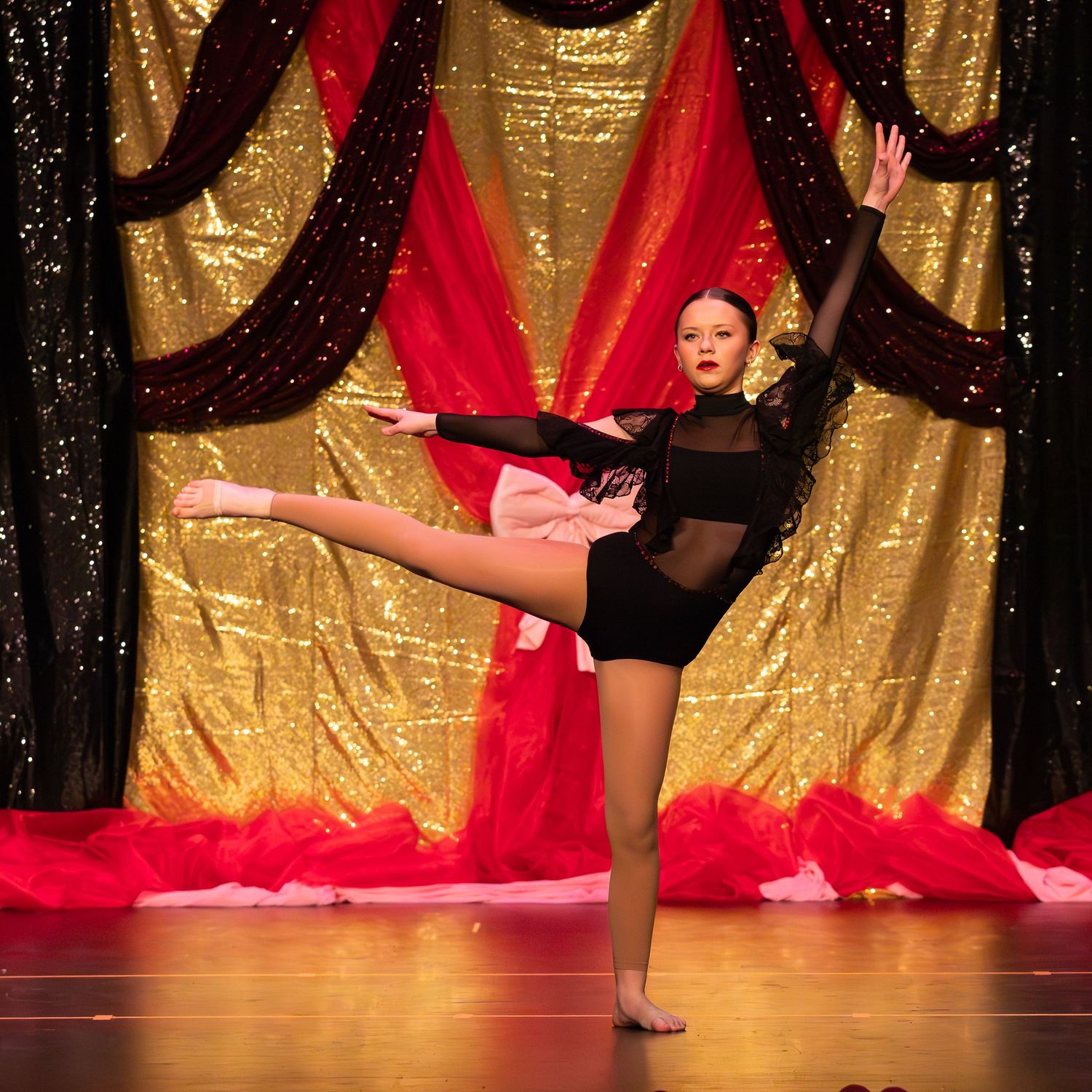A young dancer performs a high kick on stage in a black leotard against a gold sequin backdrop.