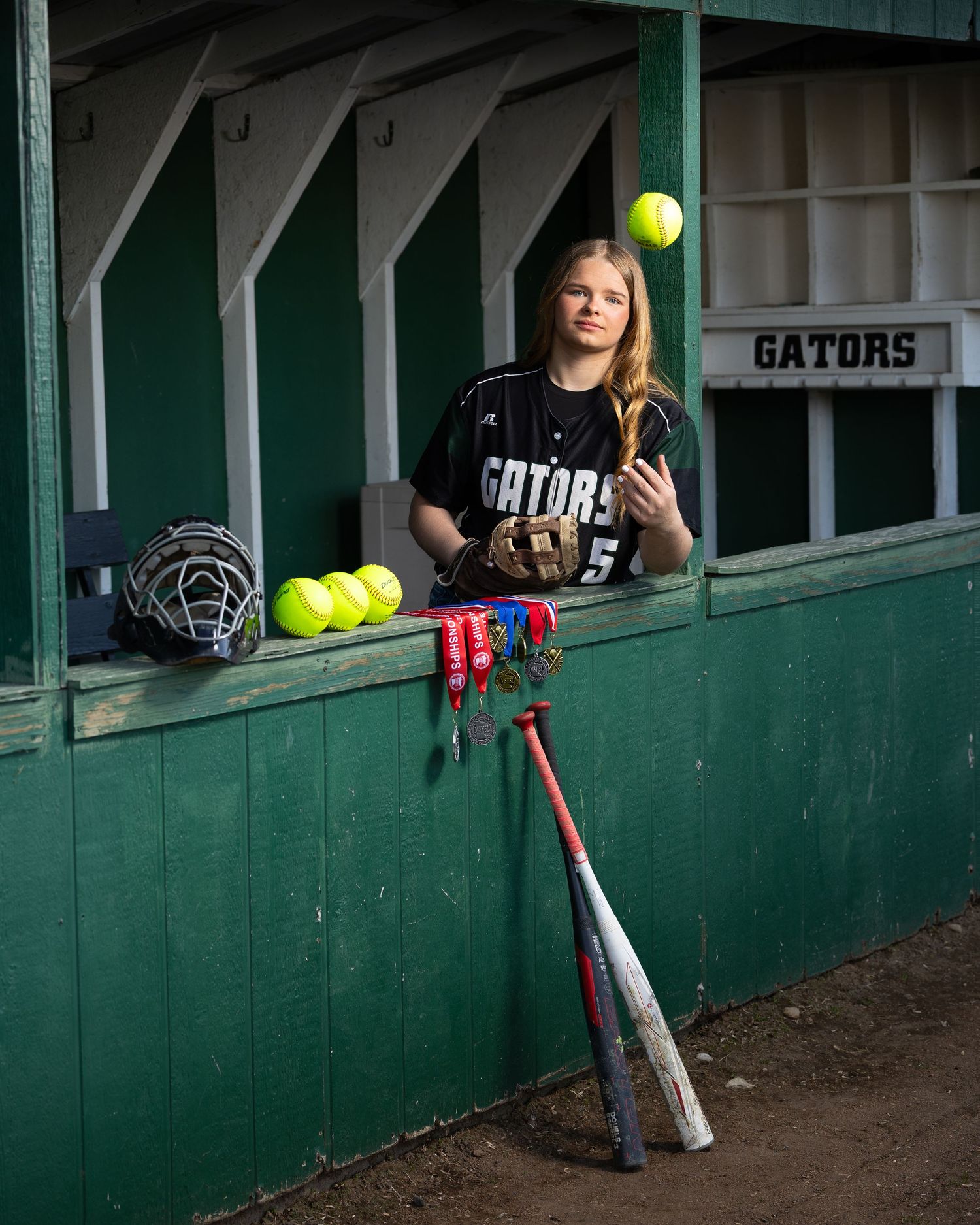 Teen girl in Gators softball jersey tosses ball in dugout with bat, glove, and medals displayed nearby.