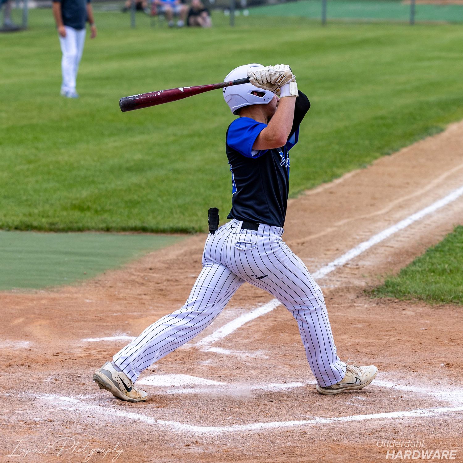 Youth baseball player in pinstripe pants swings a bat at home plate during a sunny outdoor game.