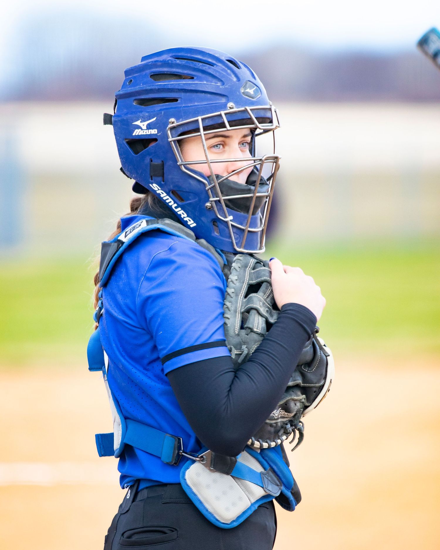 Female softball catcher in blue Mizuno helmet and chest protector holding mitt during a game.