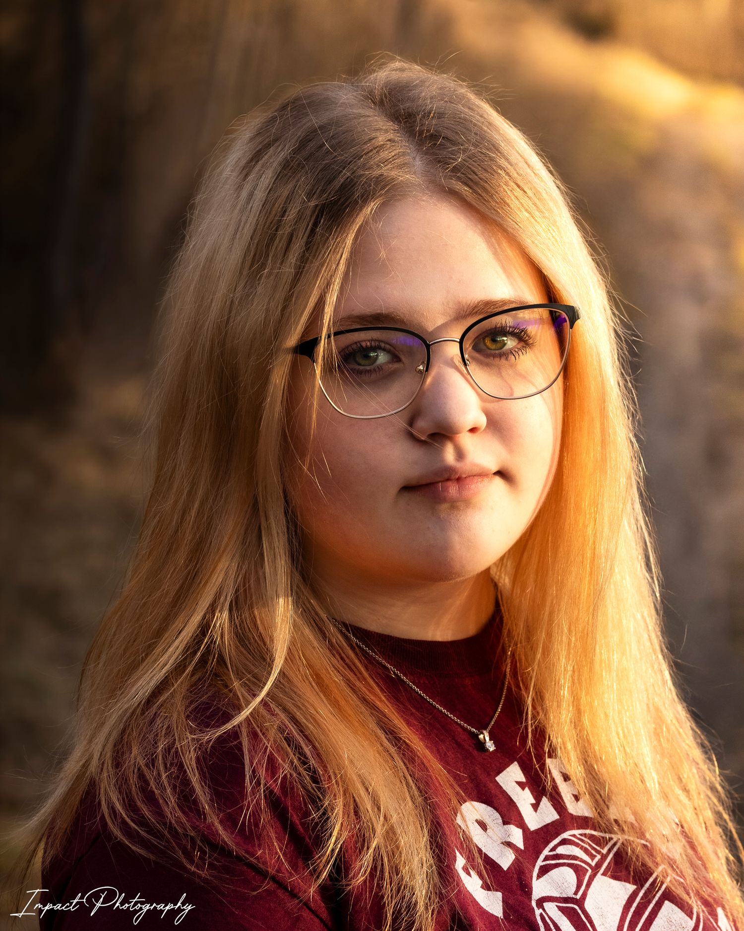 Young woman with long blonde hair and glasses poses outdoors in golden hour light wearing a maroon shirt.