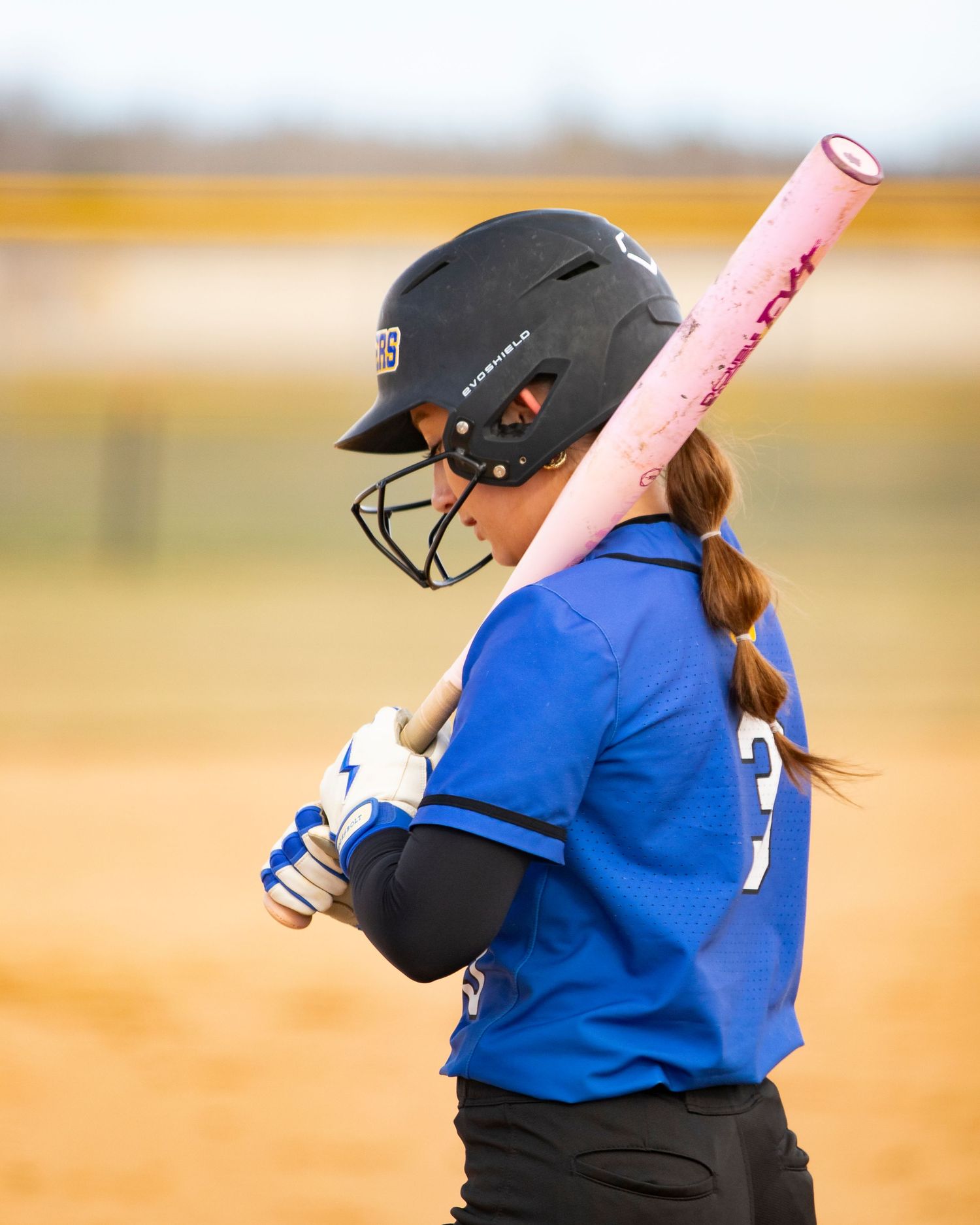 Female softball player in blue jersey holds pink bat over shoulder, wearing black helmet with face guard on dirt field.