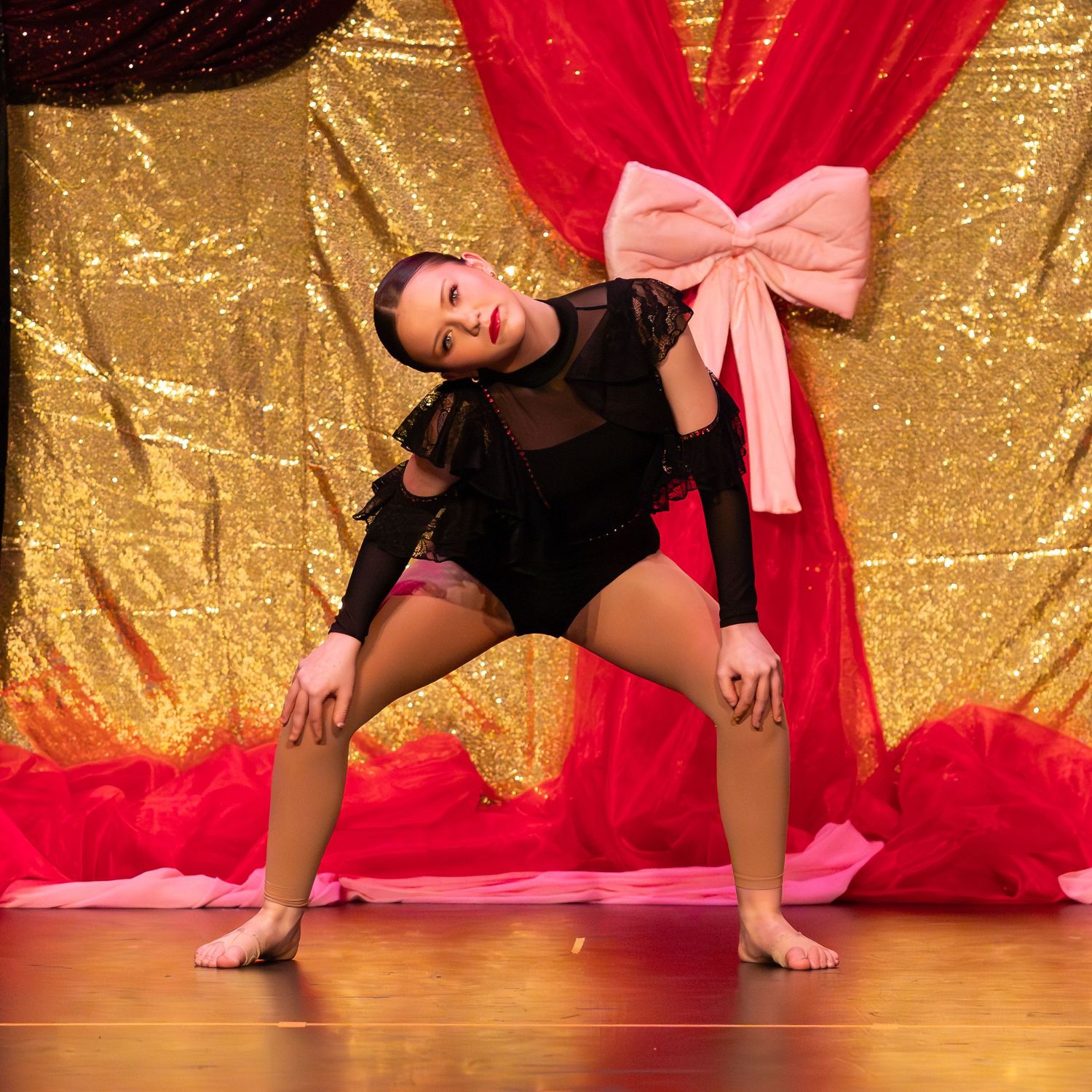 Dancer in black mesh bodysuit strikes a dramatic pose on stage with gold sequin and red drape backdrop.