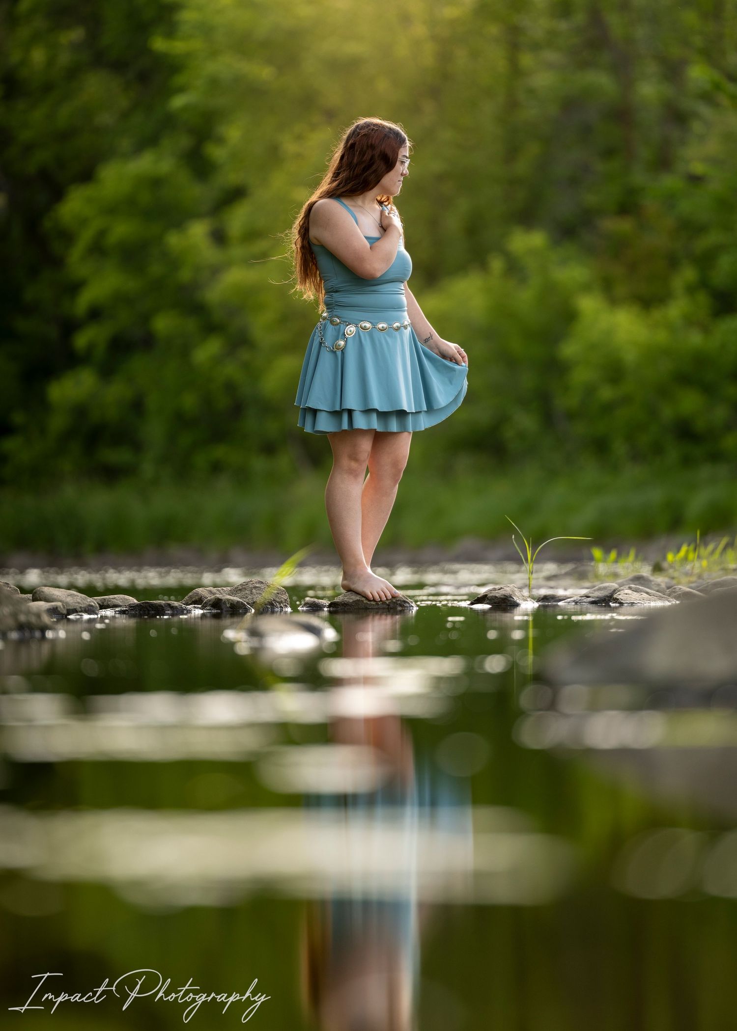 Young woman in teal dress standing barefoot on rocks in a serene stream, with lush green trees and her reflection in the water.
