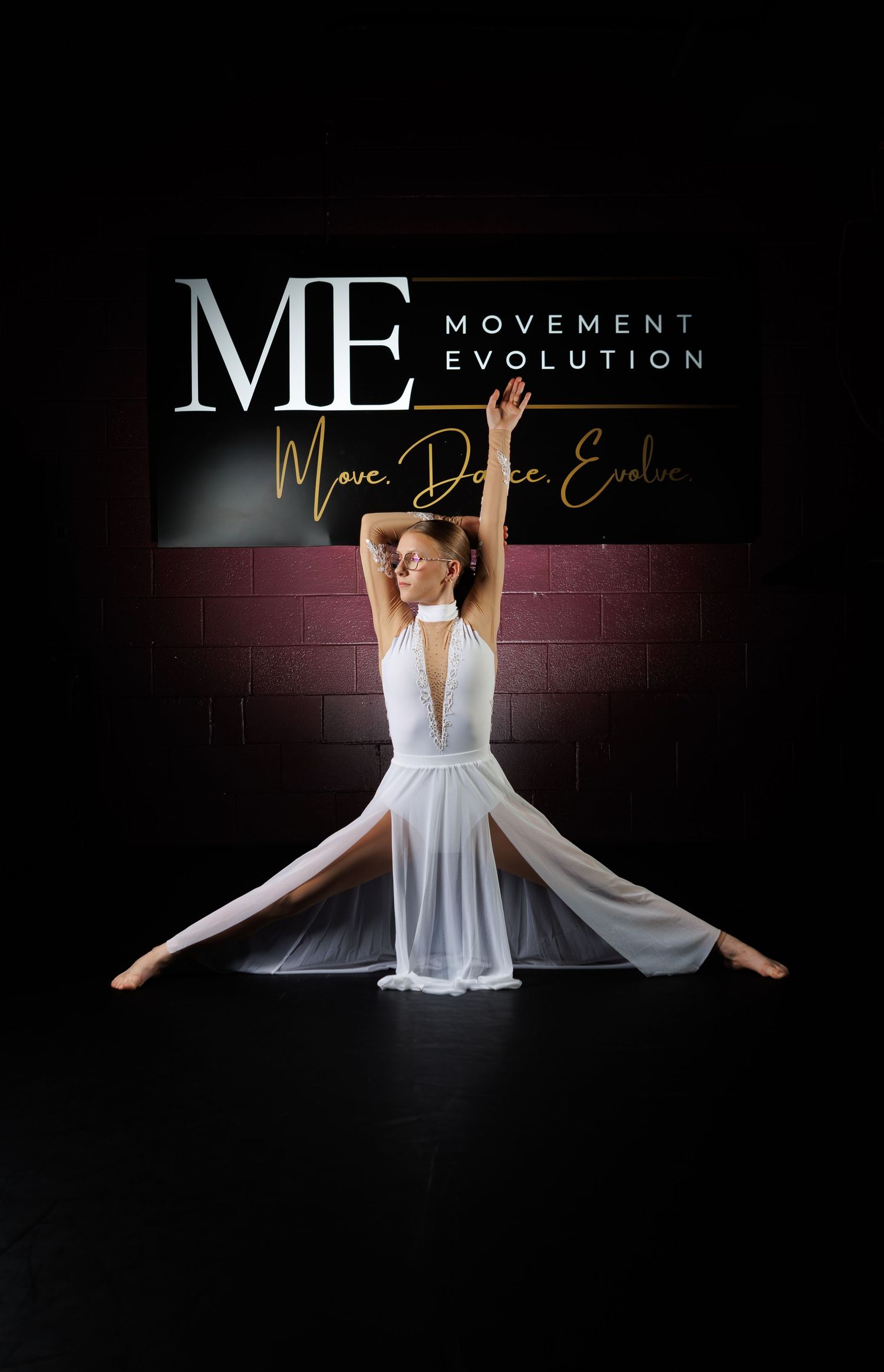 Dancer in white lyrical costume performs splits pose before Movement Evolution studio sign.