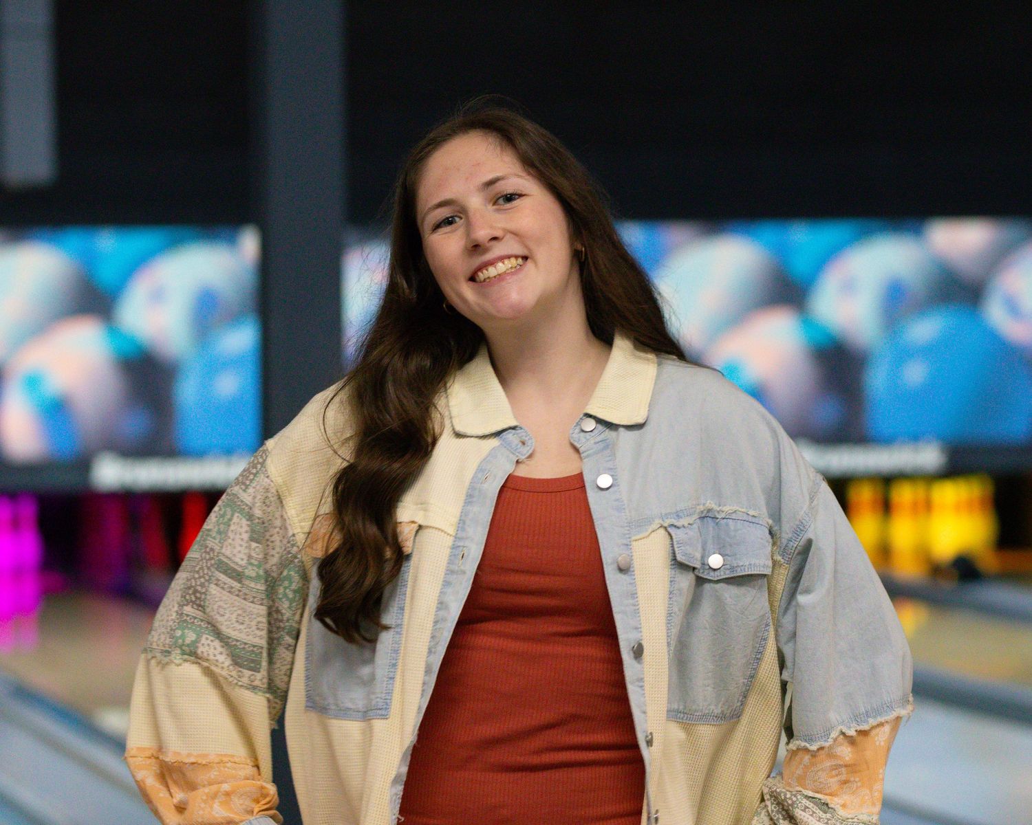 Smiling young woman in a denim jacket and red top standing in a bowling alley.