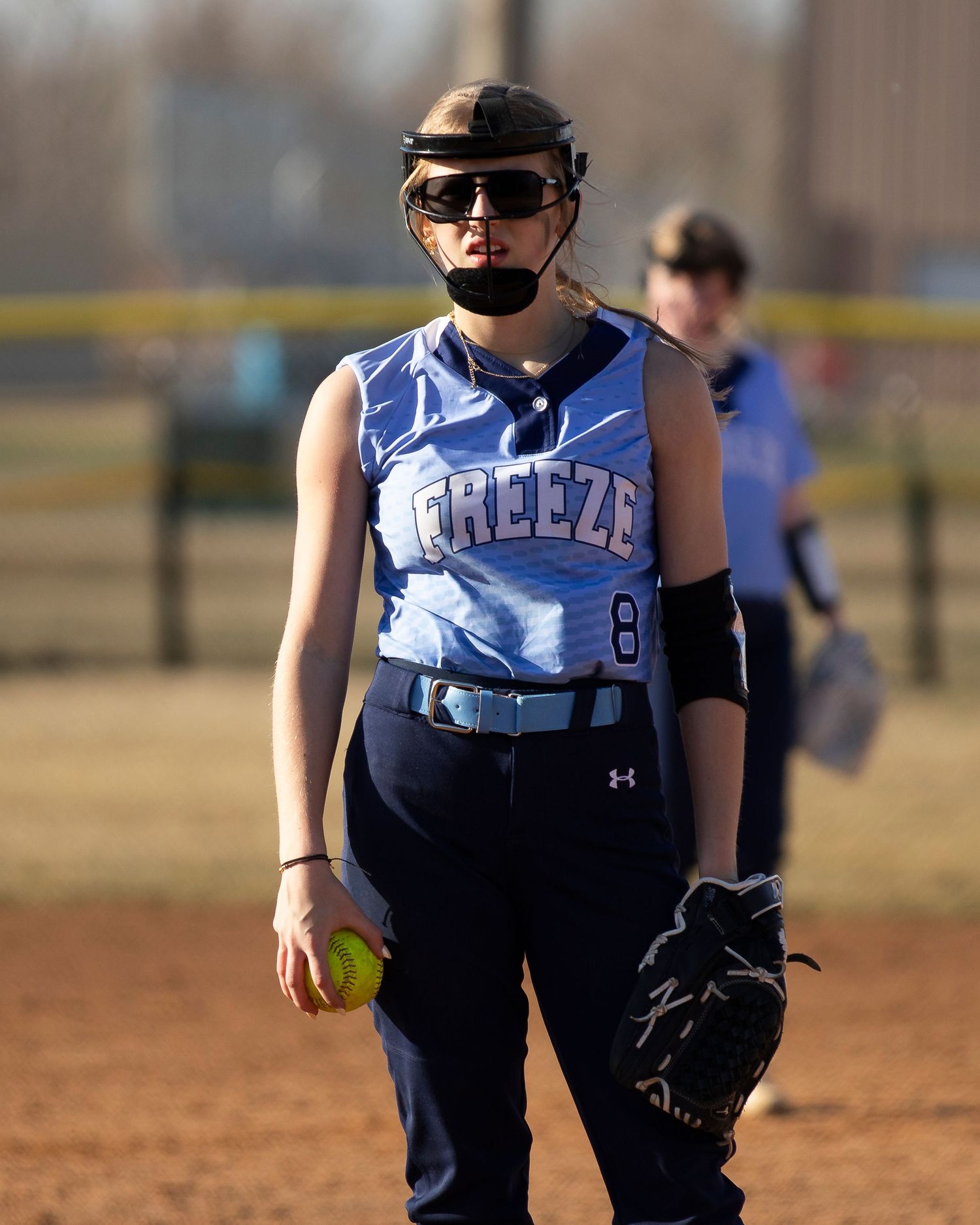 Young softball player in Freeze jersey number 8 holding a yellow ball and glove on the field.