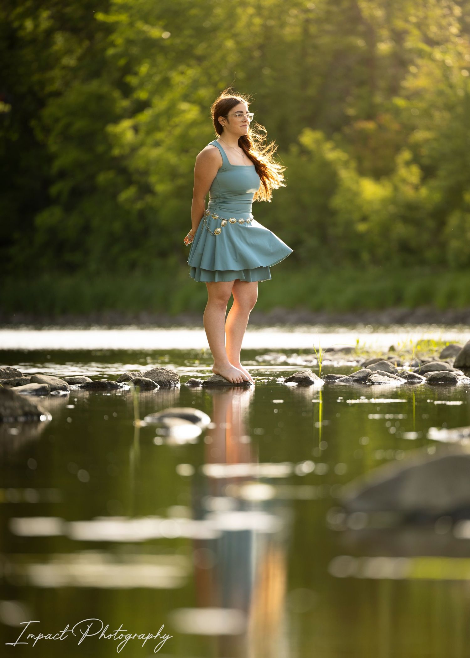 Young woman in teal dress standing on rocks in a serene reflective river, surrounded by lush green trees in golden light.