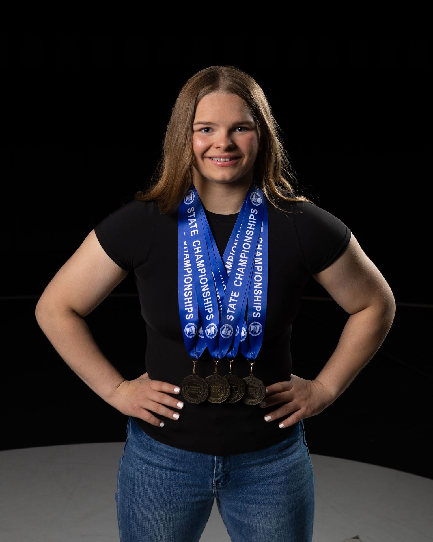 Young female athlete proudly poses with multiple state championship medals on blue ribbons, wearing a black top and jeans.