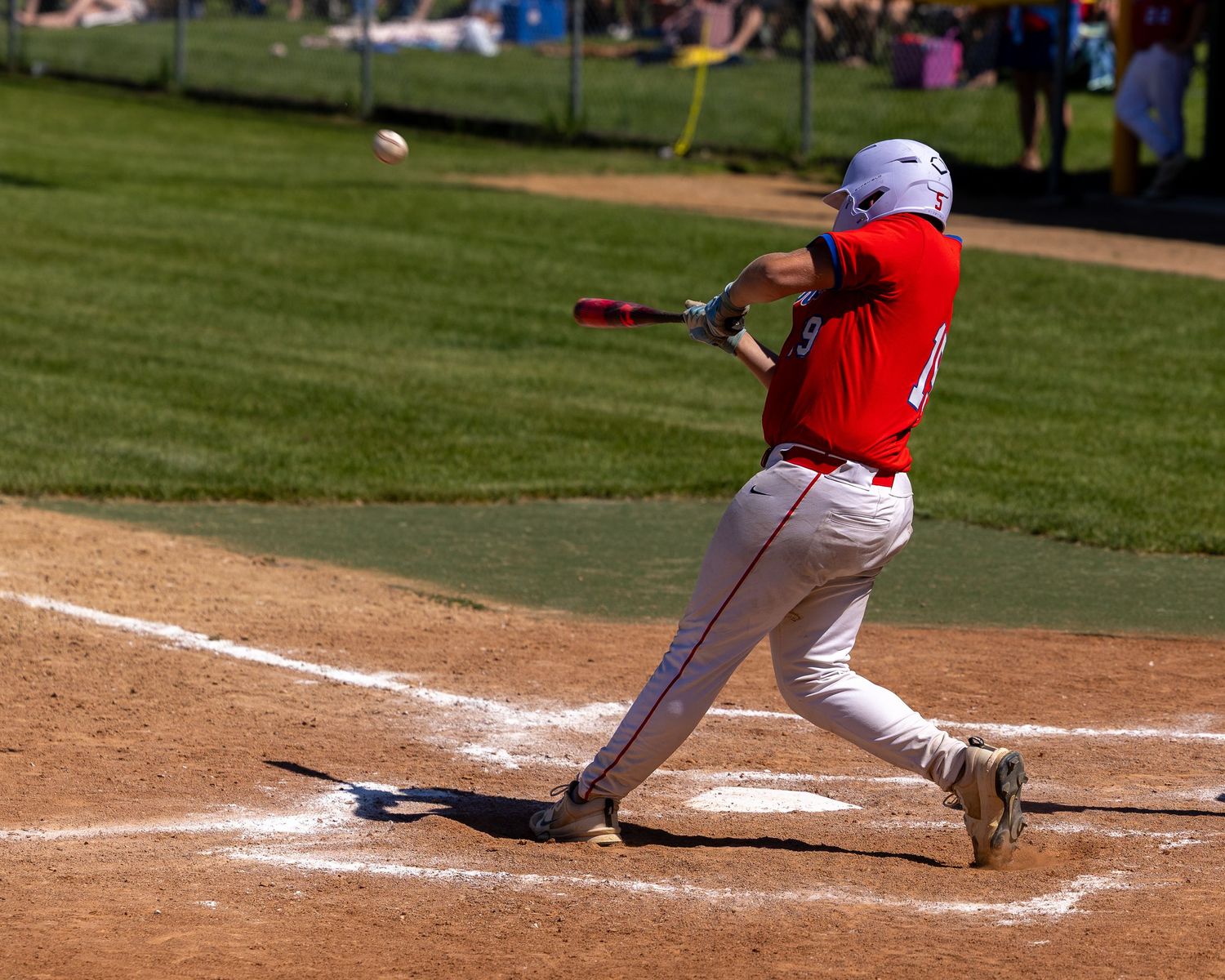 Baseball player in red uniform swinging bat at pitch during sunny outdoor game on dirt infield.
