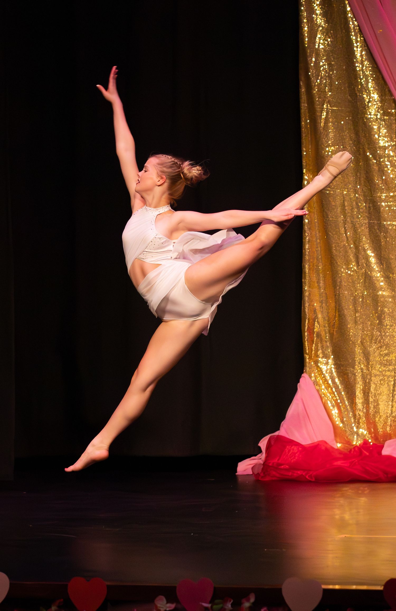 A dancer in white performs a high leg extension leap on stage with gold sequined curtains.