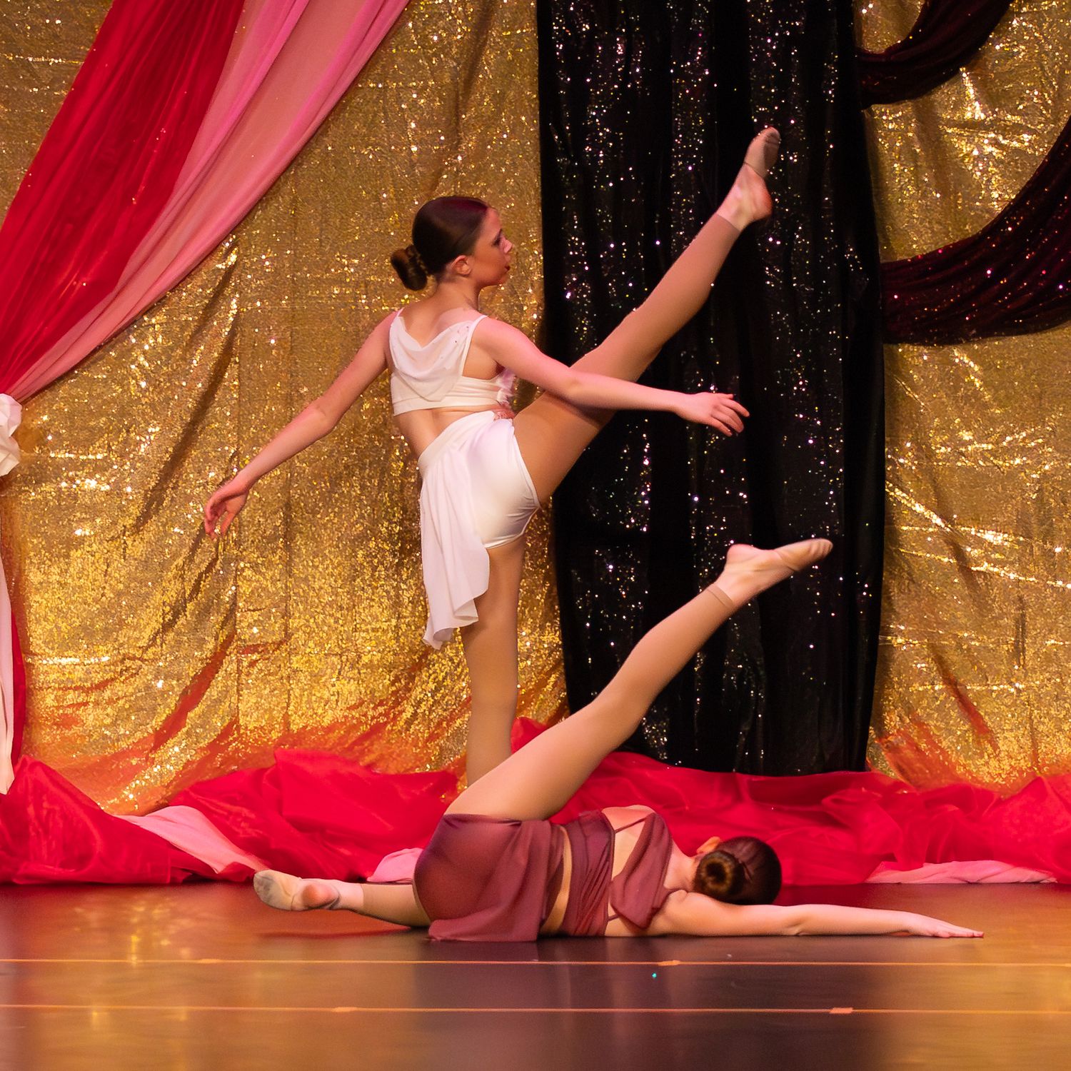 Two dancers perform contemporary ballet on stage with gold sequin backdrop, one executing a high leg extension in white costume.
