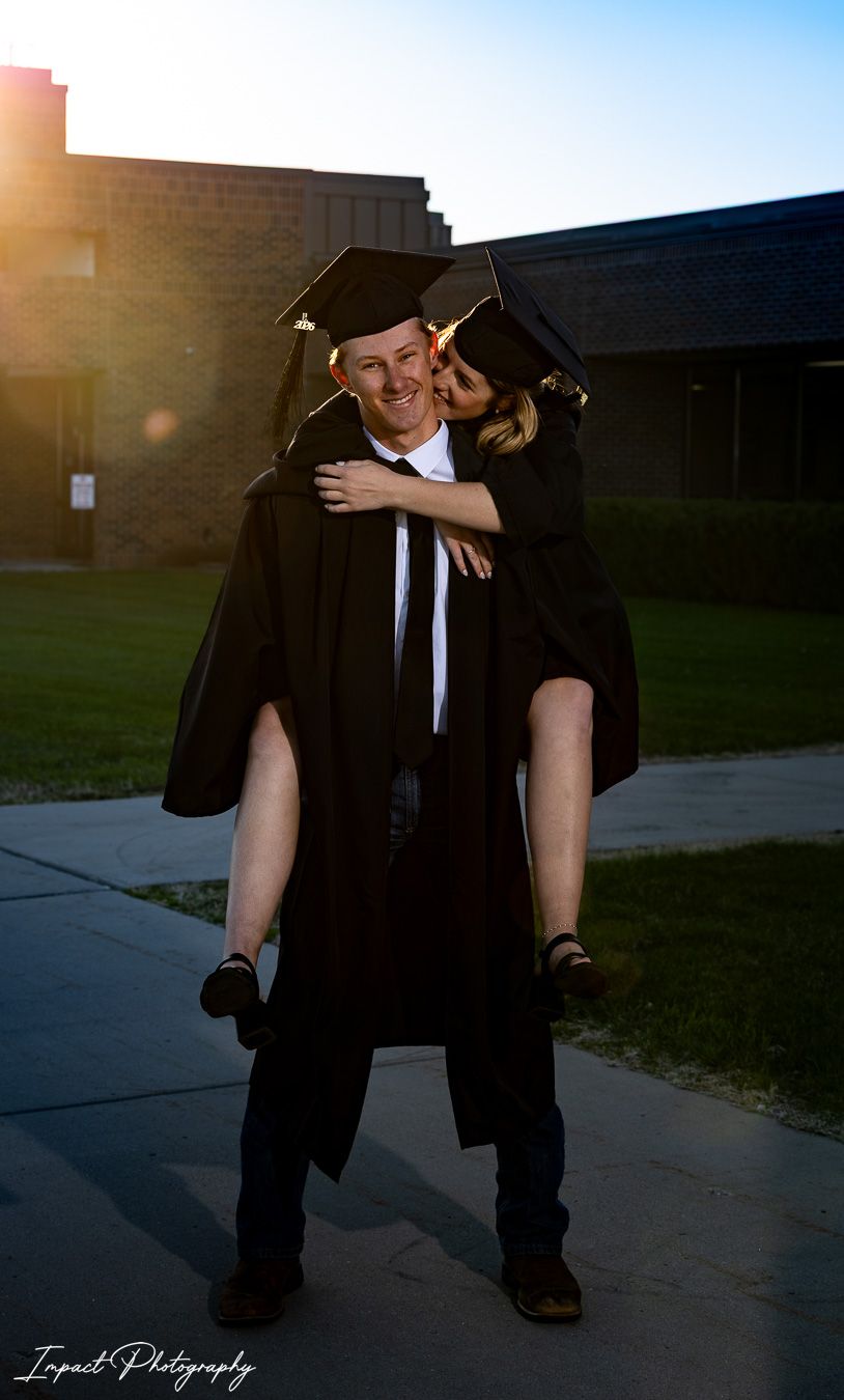 Two graduates in black caps and gowns celebrate outdoors, with one piggyback riding the other at sunset.