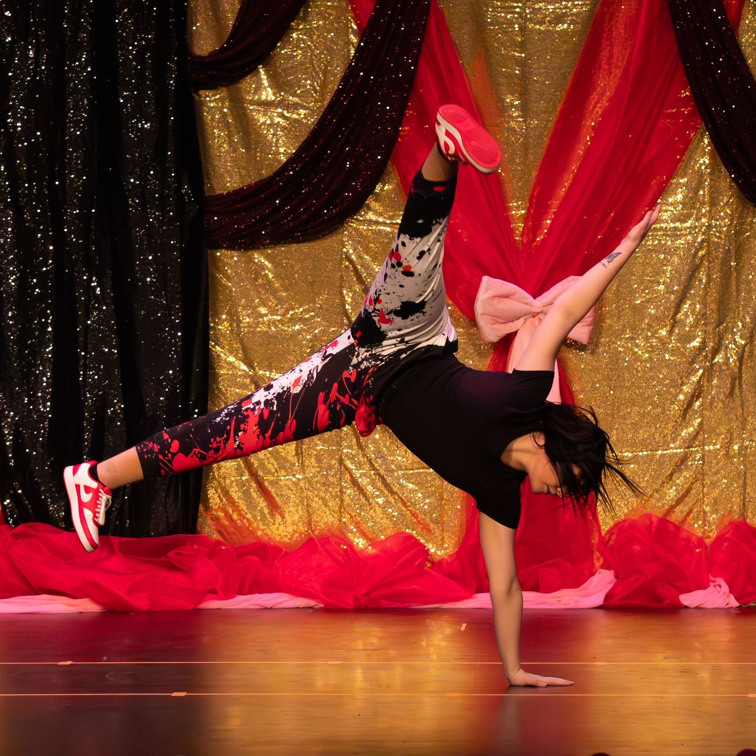 Young dancer performing a one-handed freeze breakdance move on stage with gold backdrop.