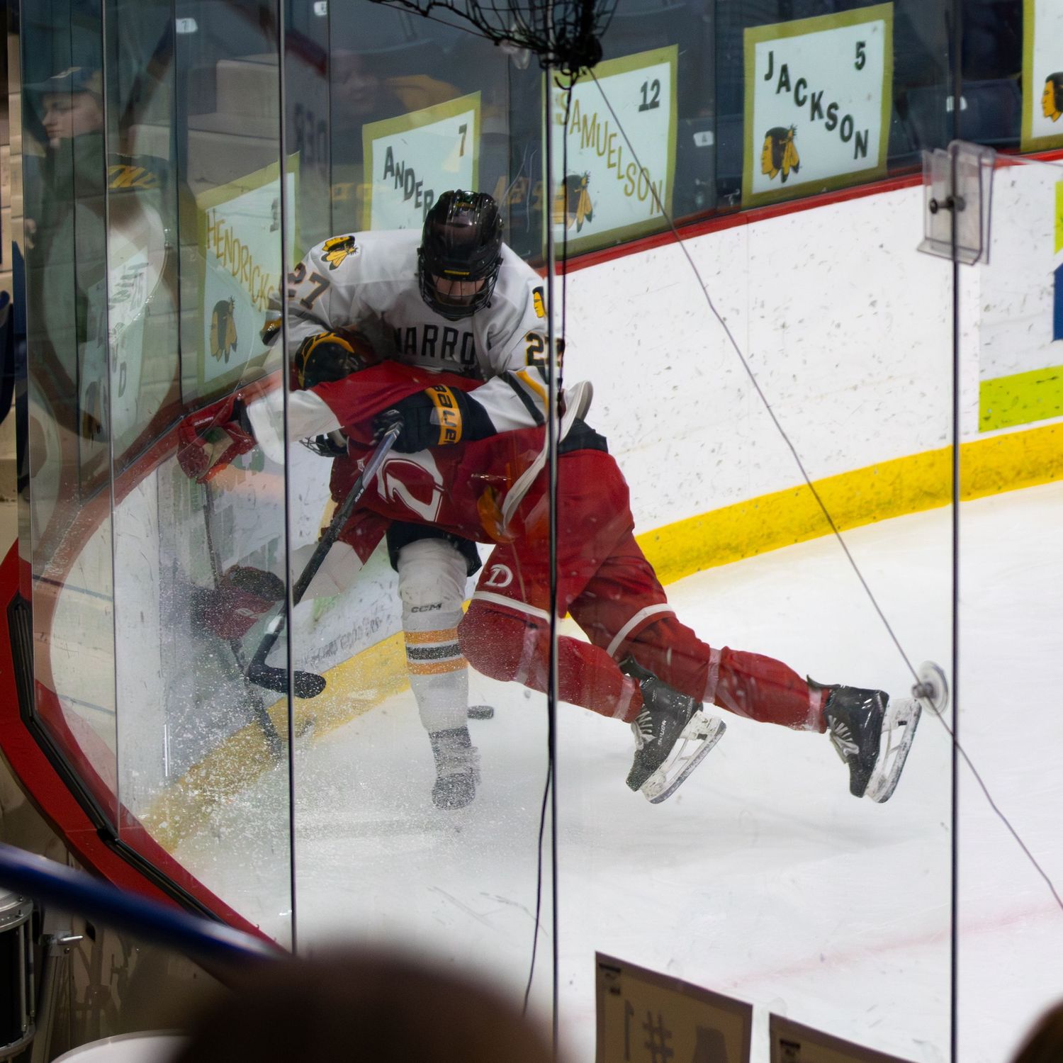 Hockey player slams opponent into glass boards during intense competitive ice hockey game action.