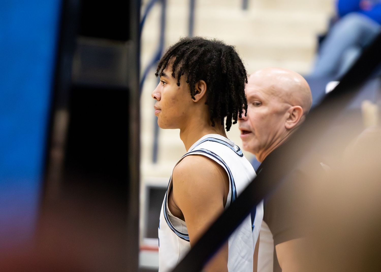 Young basketball player with dreadlocks stands beside bald coach during high school game.