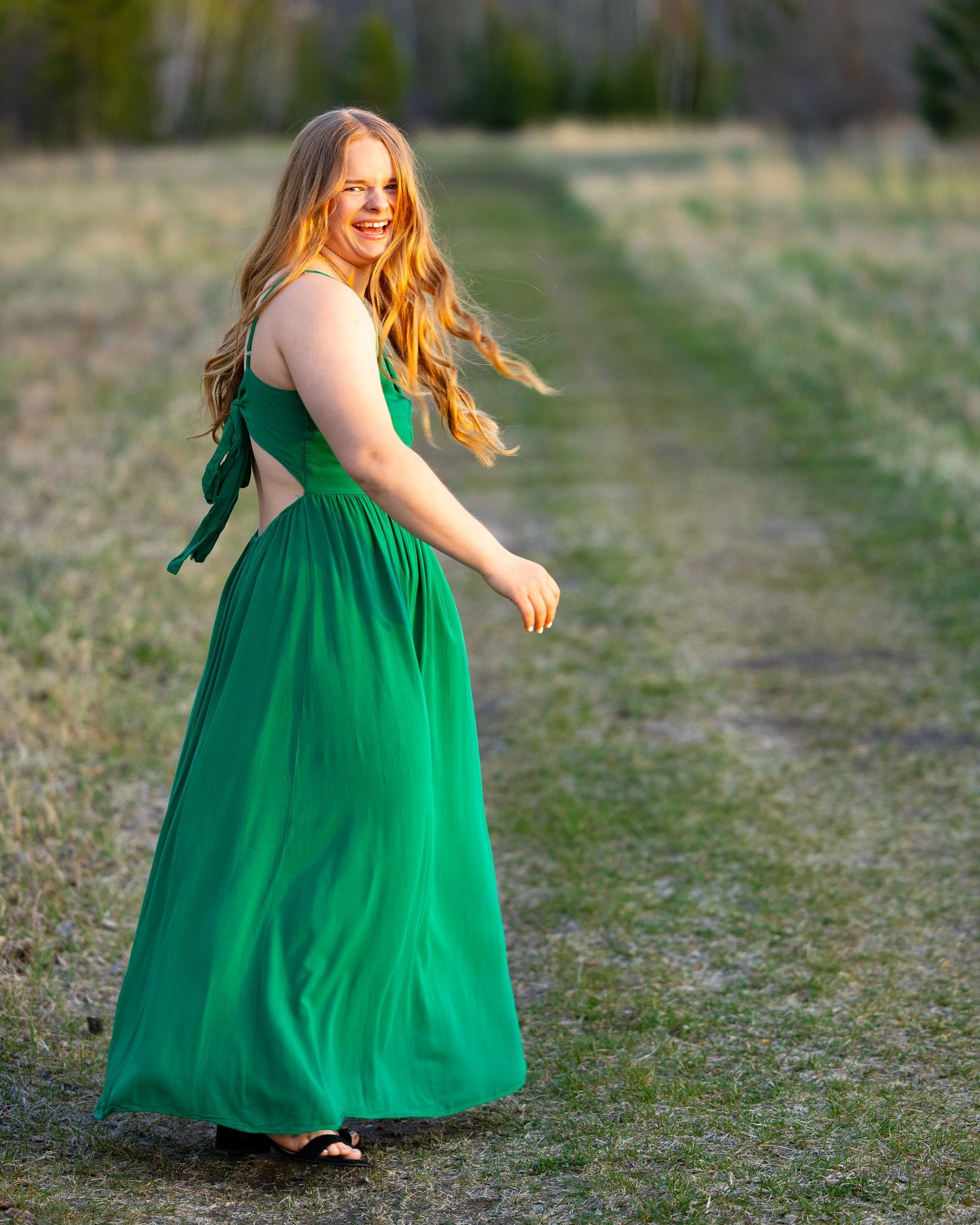 Young woman in elegant green maxi dress twirling on a rural path, smiling over her shoulder in golden hour light.
