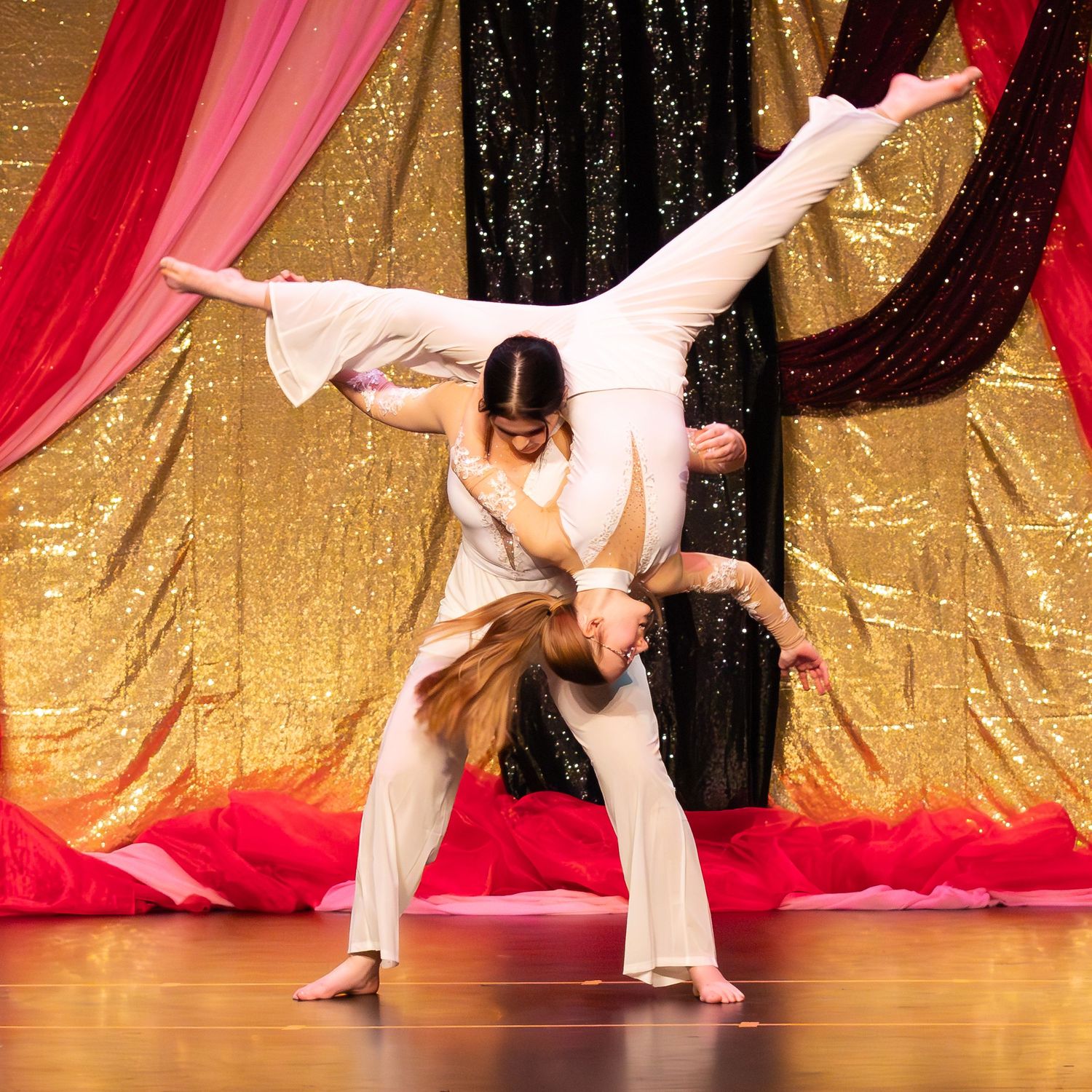 Two dancers perform an acrobatic lift on stage in white costumes with a gold sequin backdrop.