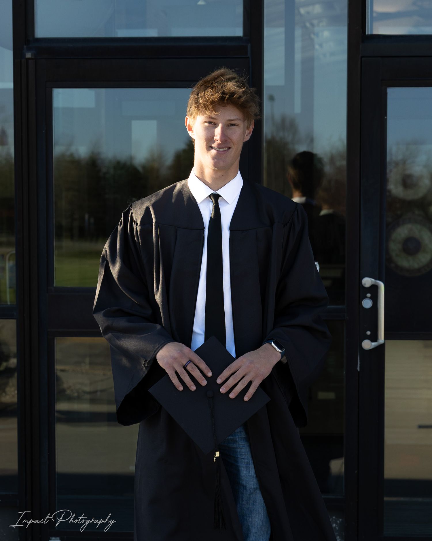 Young man in black graduation gown and tie smiling outdoors in front of glass doors on a sunny day.