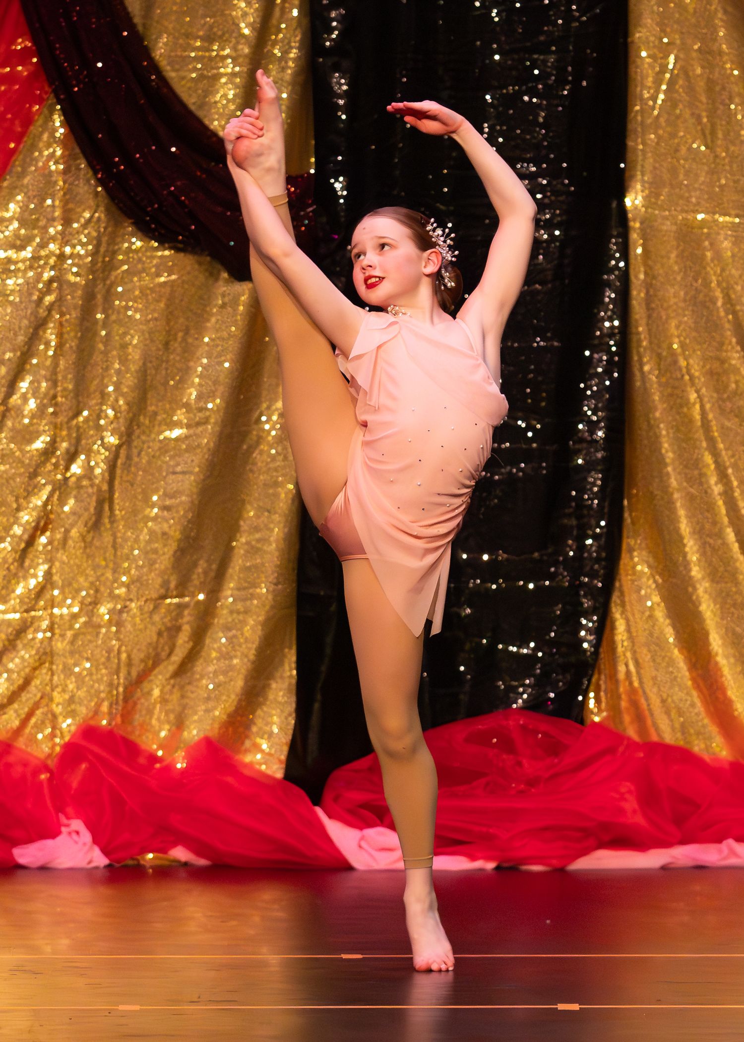 A young ballet dancer performs a high leg extension in a pink costume on a glittering gold stage.