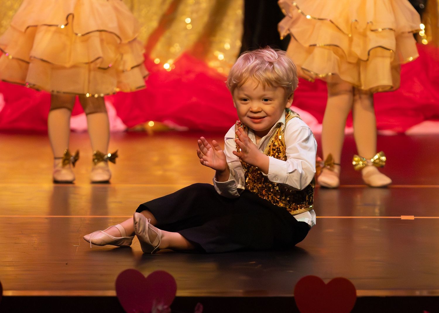 Young child in costume clapping and smiling while sitting on stage during a dance recital.