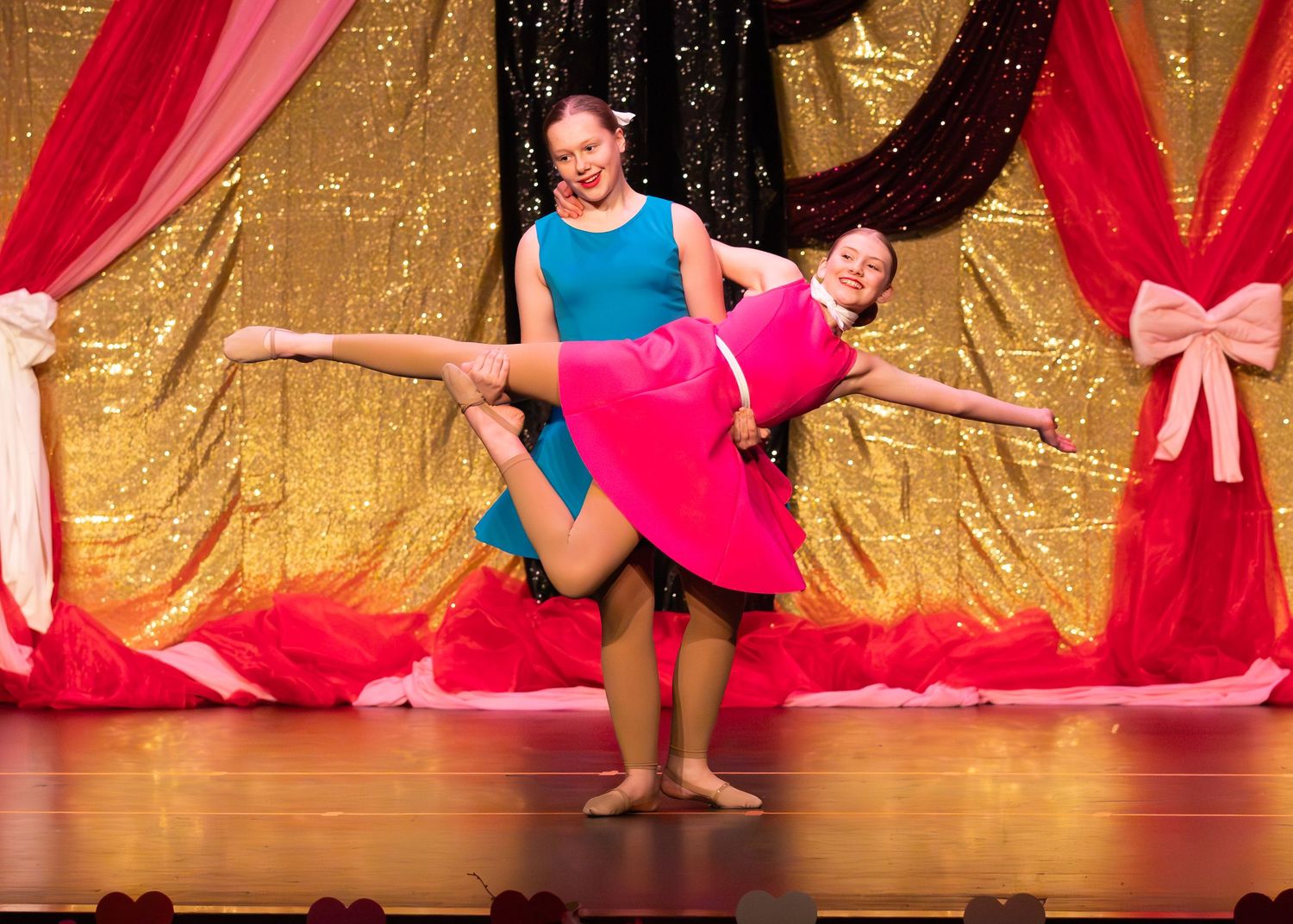 Two young dancers perform a lift on stage in colorful costumes against a gold sequin backdrop.