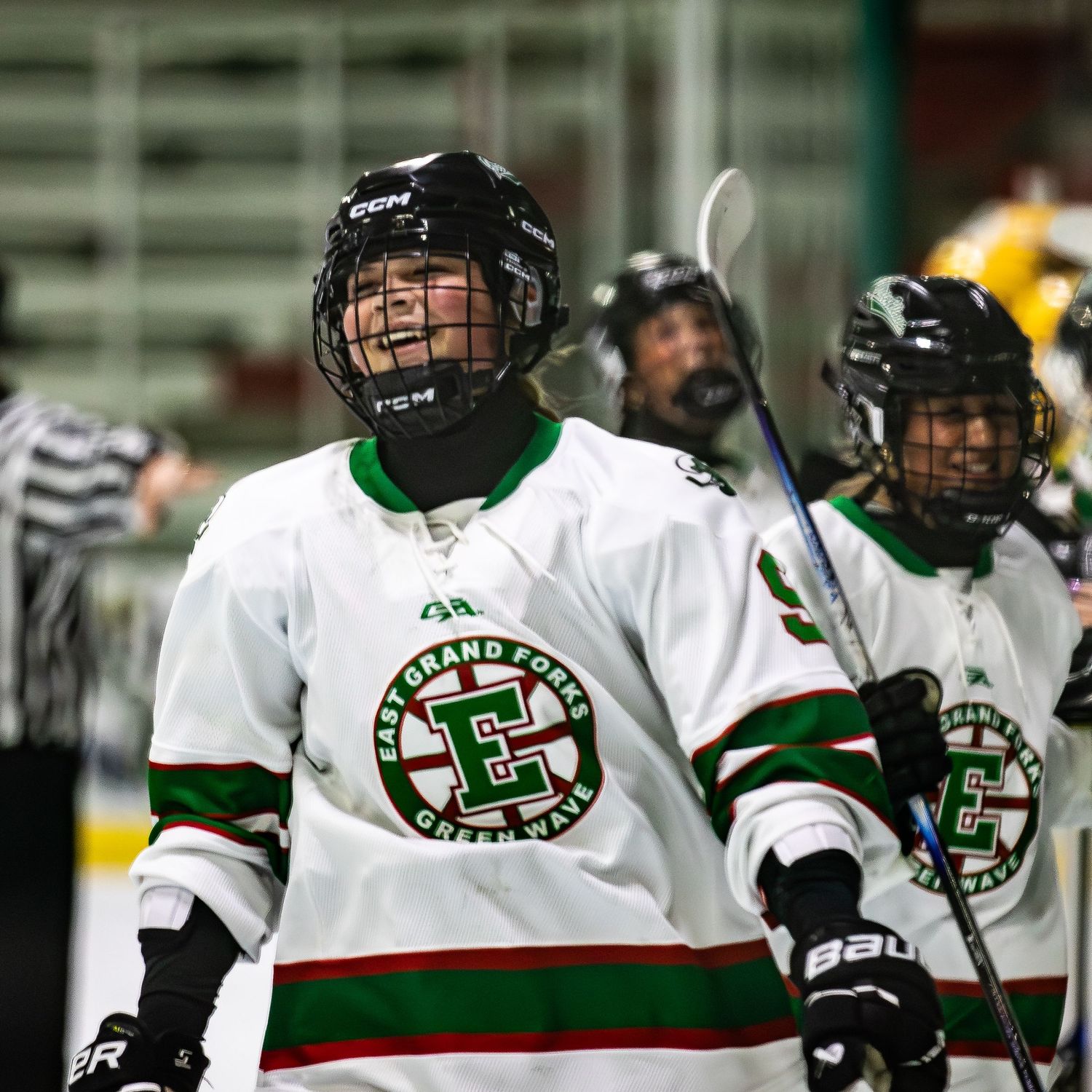 Smiling youth hockey player in white East Grand Forks Green Wave jersey celebrates on ice rink with teammates.
