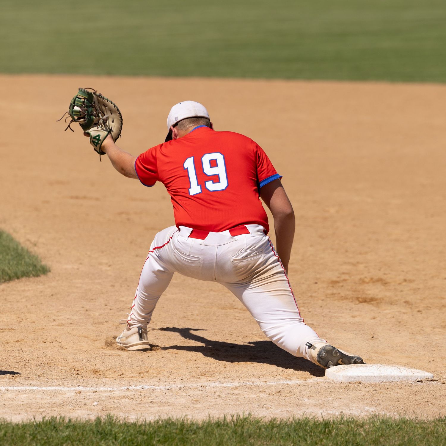 Baseball first baseman #19 in red jersey stretches to catch ball at first base during sunny day game.
