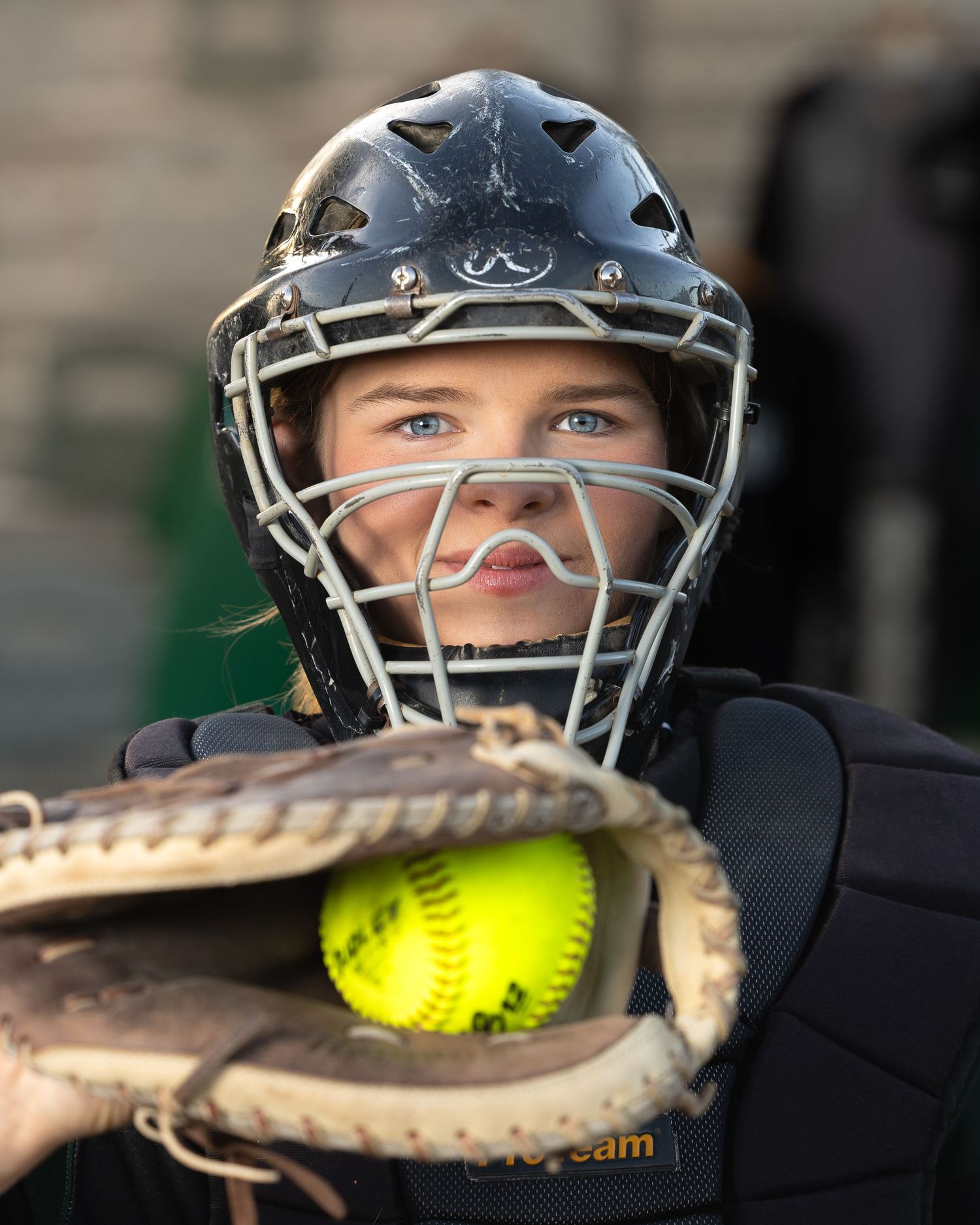 Young female softball catcher in black helmet and chest guard holds glove with yellow softball ready to play.