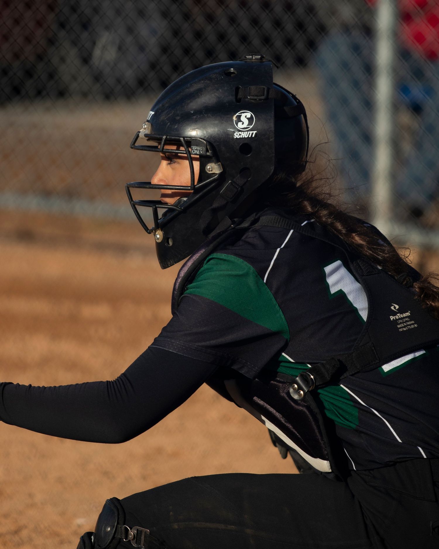 Female softball catcher in black and green uniform with Schutt helmet and face mask in ready position.