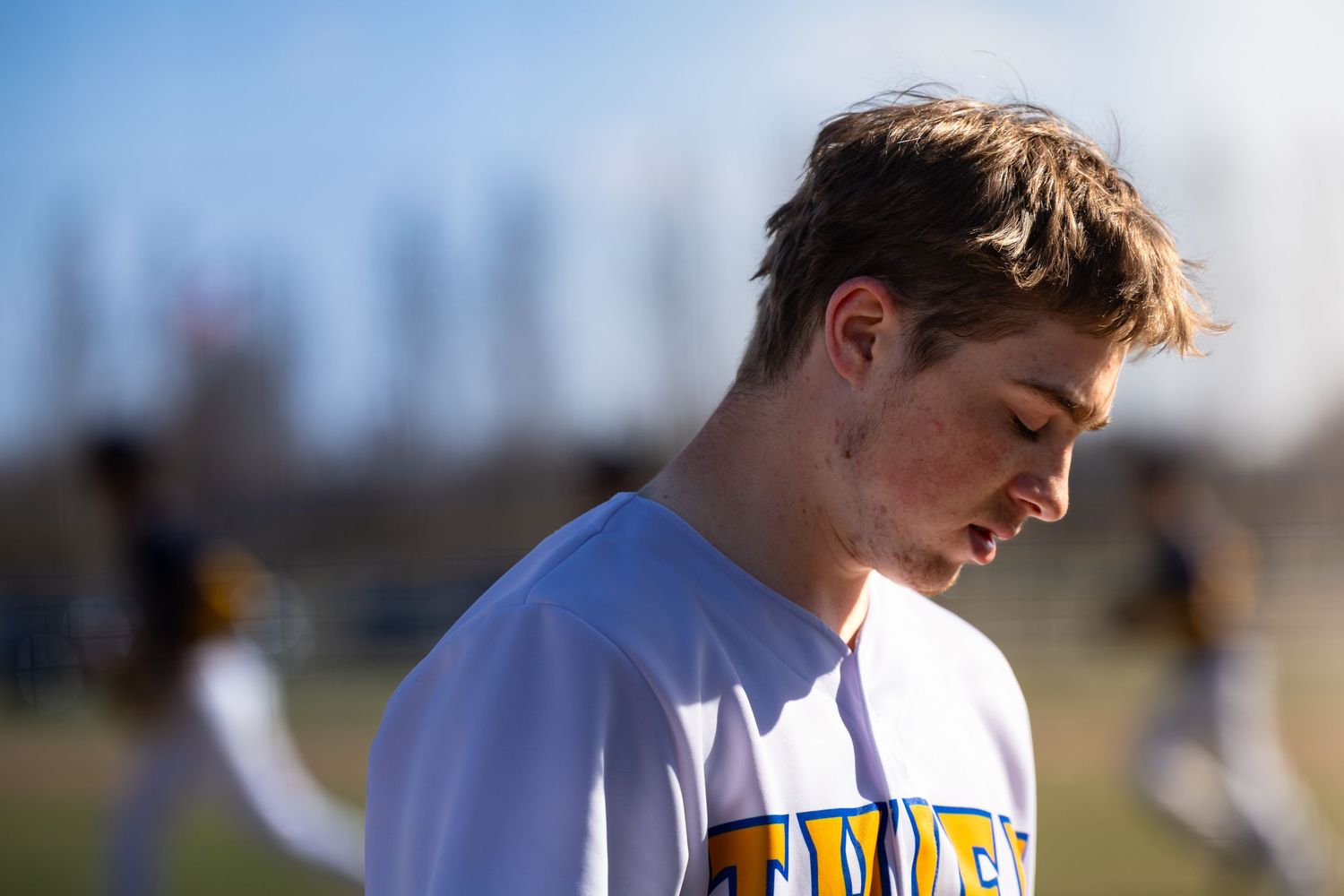 Young male athlete in white and yellow team jersey looking down on a sunny outdoor sports field.