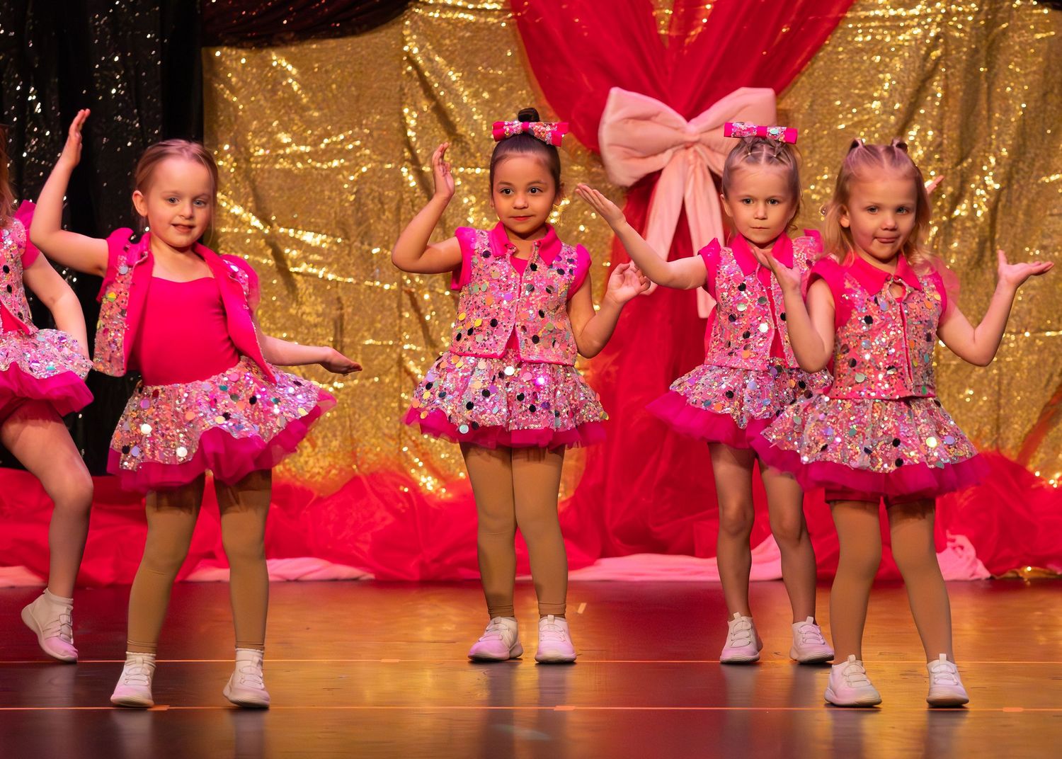 Four young girls in pink sparkly costumes dancing on stage at a children's dance recital.