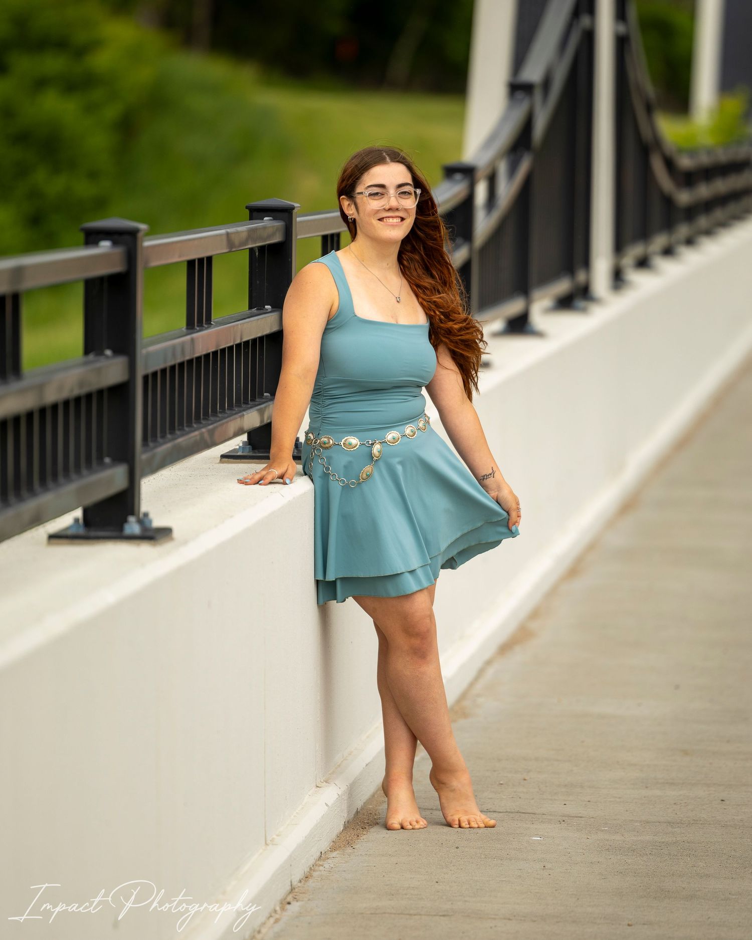 Young woman in teal dress poses barefoot on a bridge, smiling, with green trees in the background.